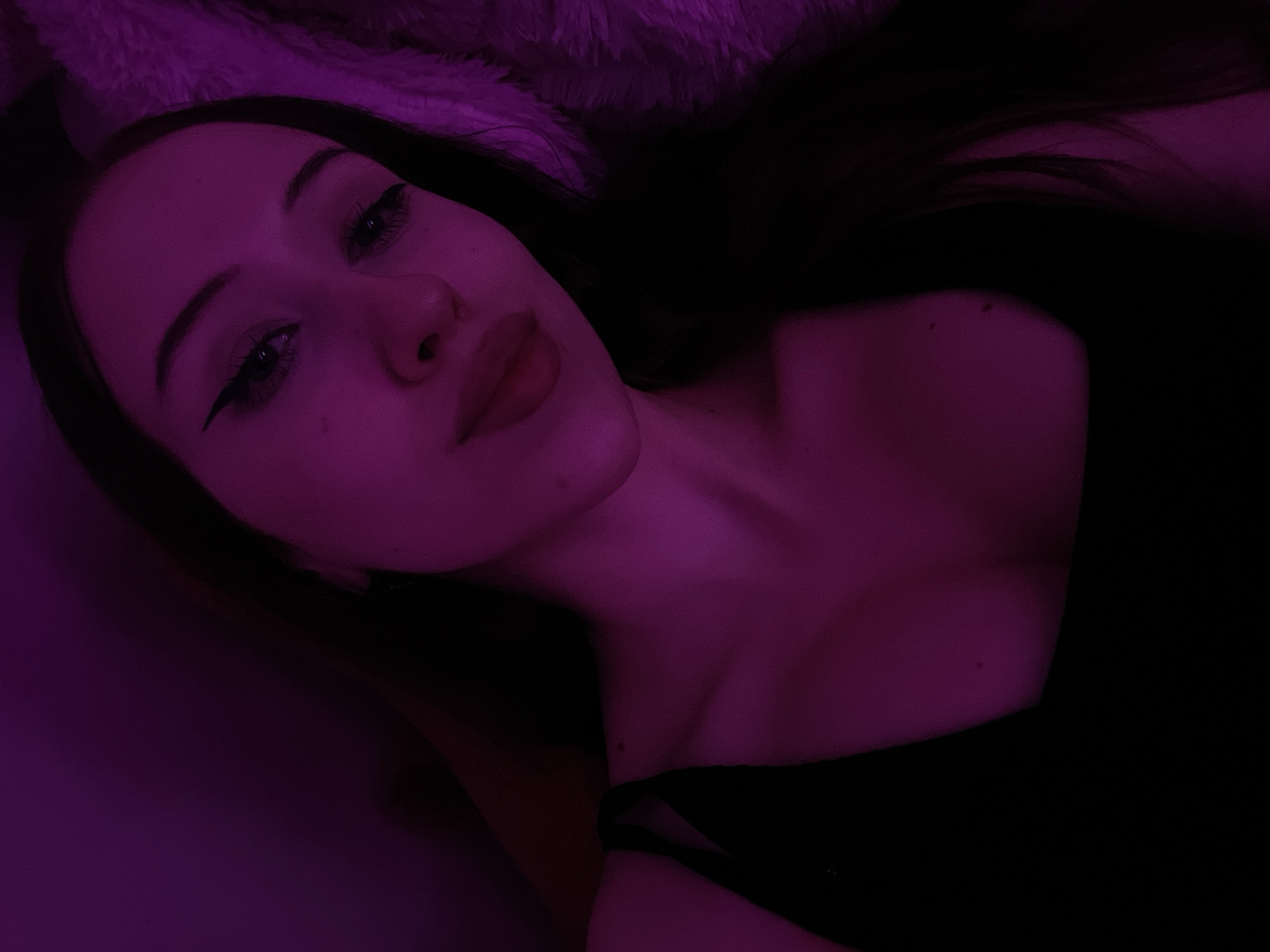 The image is a close-up, low-angle selfie of a young woman with long, dark hair. She is wearing a black tank top and has winged eyeliner. The scene is illuminated by a purple light, casting a strong hue over her skin and hair. The background is dark and slightly blurred, with a textured surface visible on the right side. There are no discernible elements that indicate a specific location within Votkinsk, Russia. The image focuses on the woman's face and upper body.