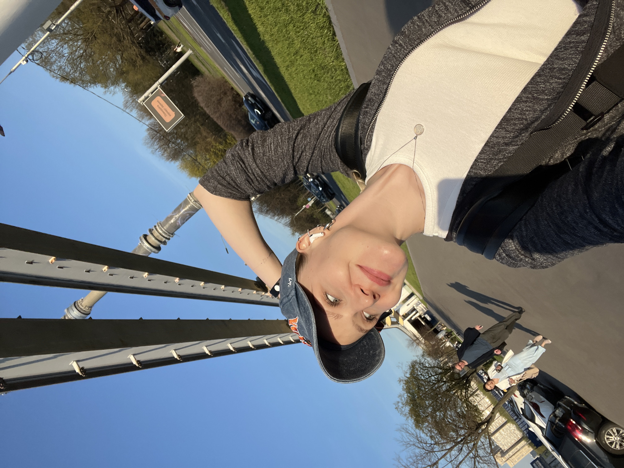 The image depicts a woman in Khimki, Russia, likely taking a selfie. She is wearing a baseball cap, a white shirt, and a gray zip-up jacket. She also has a black shoulder bag. In the background are other people on a pathway with cars parked to their right. Above her is the supports for a light rail line. A road is visible further in the background and a sign is located on the side of the road. The sky is clear and blue.