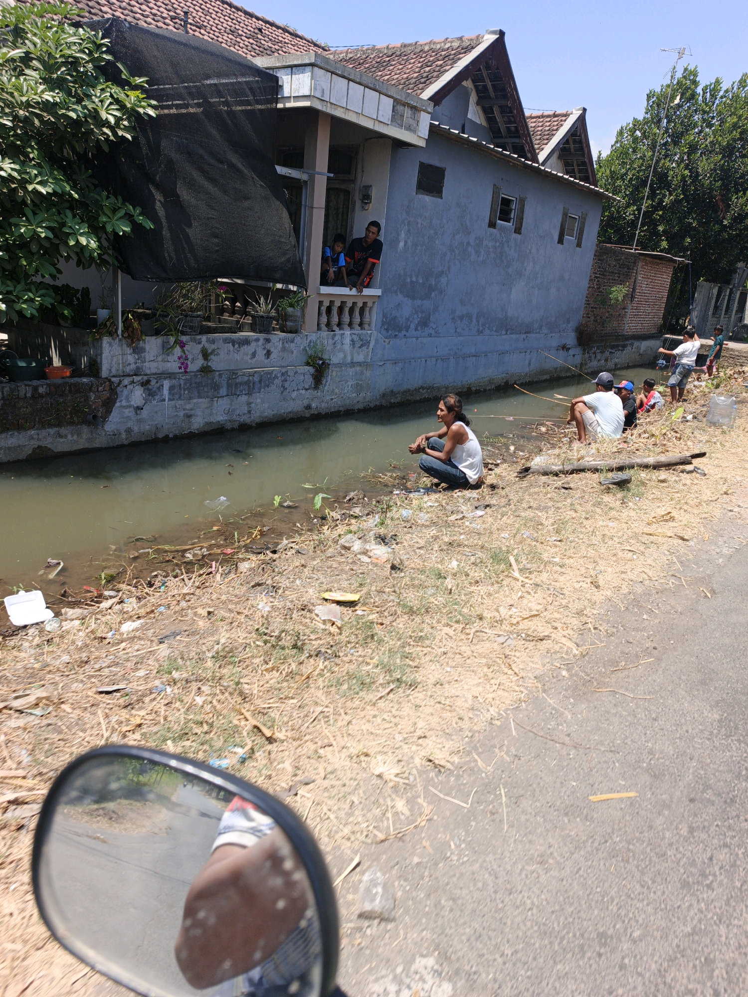A side-view mirror of a motorcycle shows a reflection of a person's arm and torso, and a rural road with dry grass and debris on the side. In the background, a group of people are fishing in a narrow, murky canal. There are several houses with red tile roofs along the canal. The houses appear to be made of concrete and have balconies. The environment is sunny with a clear blue sky. The canal bank is littered with trash. The people are casually dressed, and some are sitting on the ground while others are standing. One person on the balcony of a house is looking down at the people fishing.