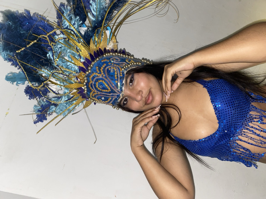 A woman poses wearing a vibrant blue sequined top with a fringed hem and a large, elaborate blue and gold feathered headdress. The headdress features intricate beaded designs and extends upwards with a variety of textures and lengths of feathers and decorative strands. She's lightly made up, gazing at the camera with her hands near her face. The background is plain white, highlighting the colorful costume.