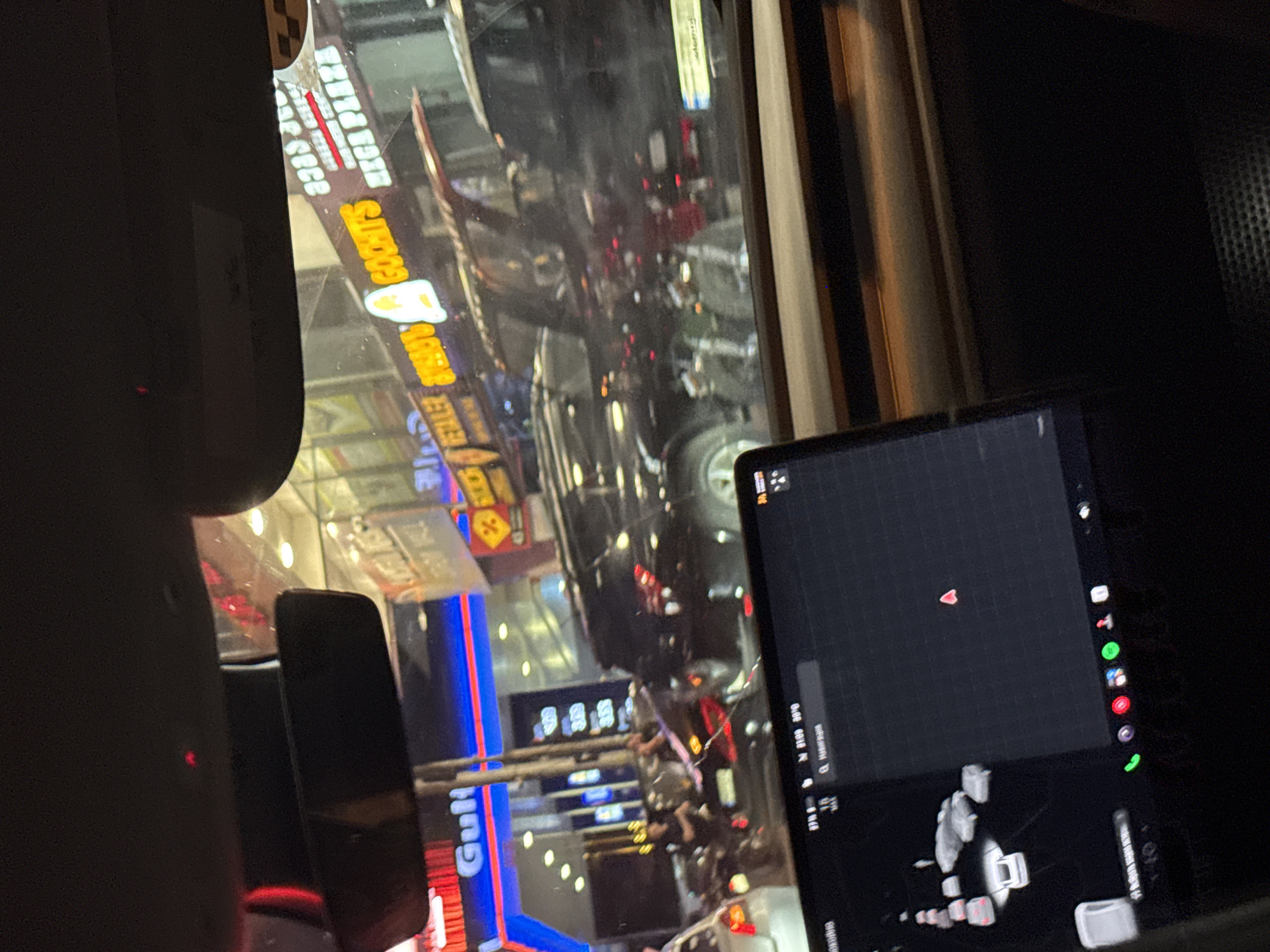 The image depicts a nighttime scene viewed from inside a vehicle in Tbilisi, Georgia, likely at a gas station. In the foreground, a car's dashboard display shows a map and various icons. Beyond the dashboard, the windshield reveals a busy street with multiple cars, including dark-colored SUVs and a white sedan. Several illuminated signs are visible, one prominently displaying "Gulf" with red and blue accents, suggesting a Gulf Oil gas station. Another sign reads "GOCHIT'S" in yellow lettering. Neon lights and reflections on the wet pavement indicate a bustling urban environment. The overall impression is one of a typical evening in a city, with traffic, commercial activity, and vehicle presence.