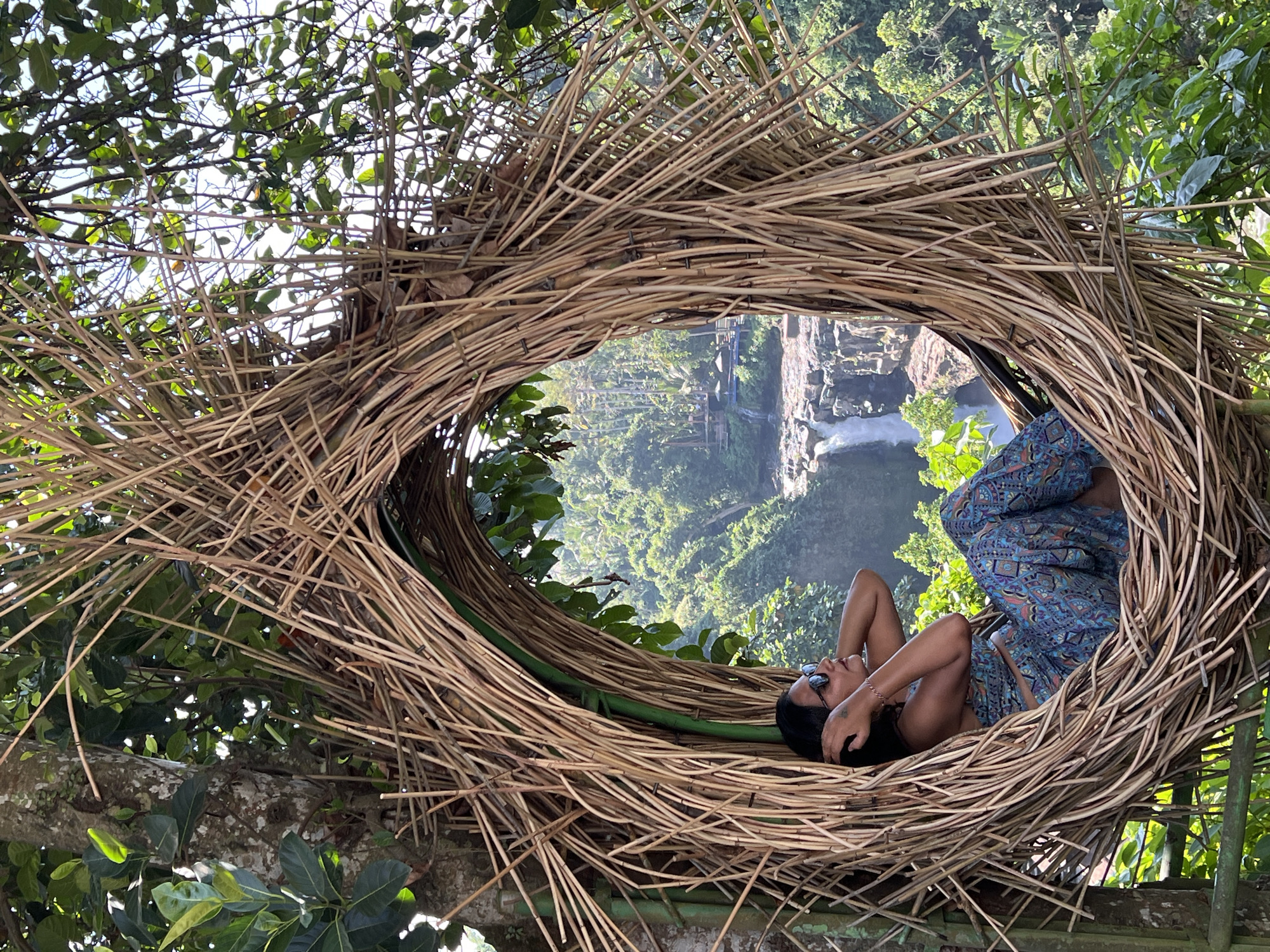 In Sukawati, Indonesia, a woman is seated in a large, nest-like structure made of woven branches. She is wearing a patterned blue and gold outfit and sunglasses, with her hands behind her head. The nest is positioned outdoors, with lush green foliage and a waterfall visible in the background. The scene appears to be a tourist attraction or scenic viewpoint.