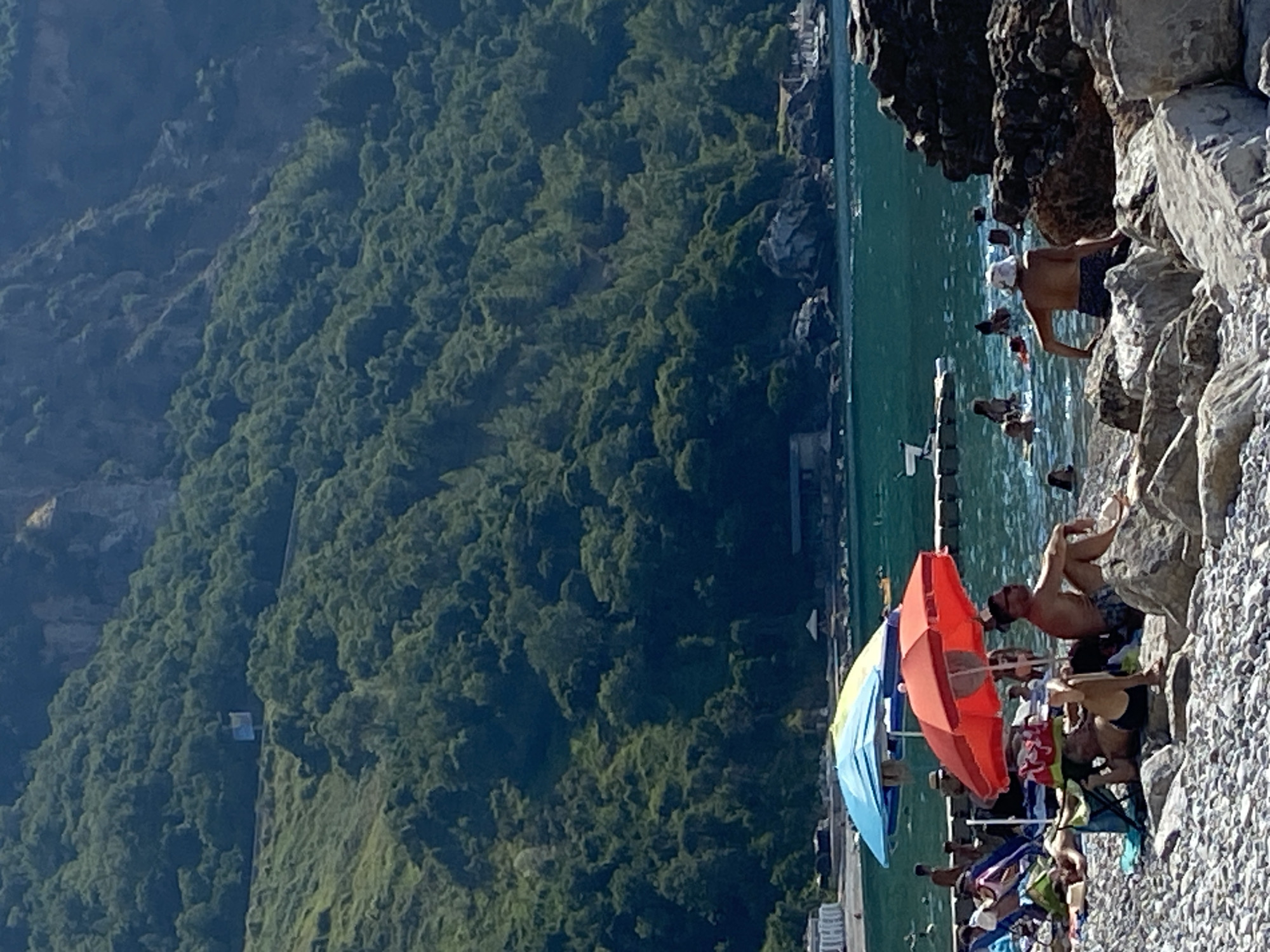 This is a full-frame, vertical, eye-level, outdoor, daytime, slightly blurry, full shot photograph taken on a sunny day at a pebbly beach in Prijevor, Montenegro. The image depicts a beach scene with a body of water and a lush green mountainside in the background. In the foreground, several people are relaxing on the beach. A red umbrella is open and positioned over two individuals sitting on rocks. Another umbrella, blue and yellow, is open behind the red one. A man in a white hat is sitting on rocks and looking out at the water, with other people swimming and wading in the turquoise sea. The water is calm, with small waves and a floating platform with a structure on it. The mountainside is densely covered with trees, and a road is visible winding along its base. Buildings and structures are partially visible along the coastline. The overall impression is of a peaceful and scenic coastal environment.