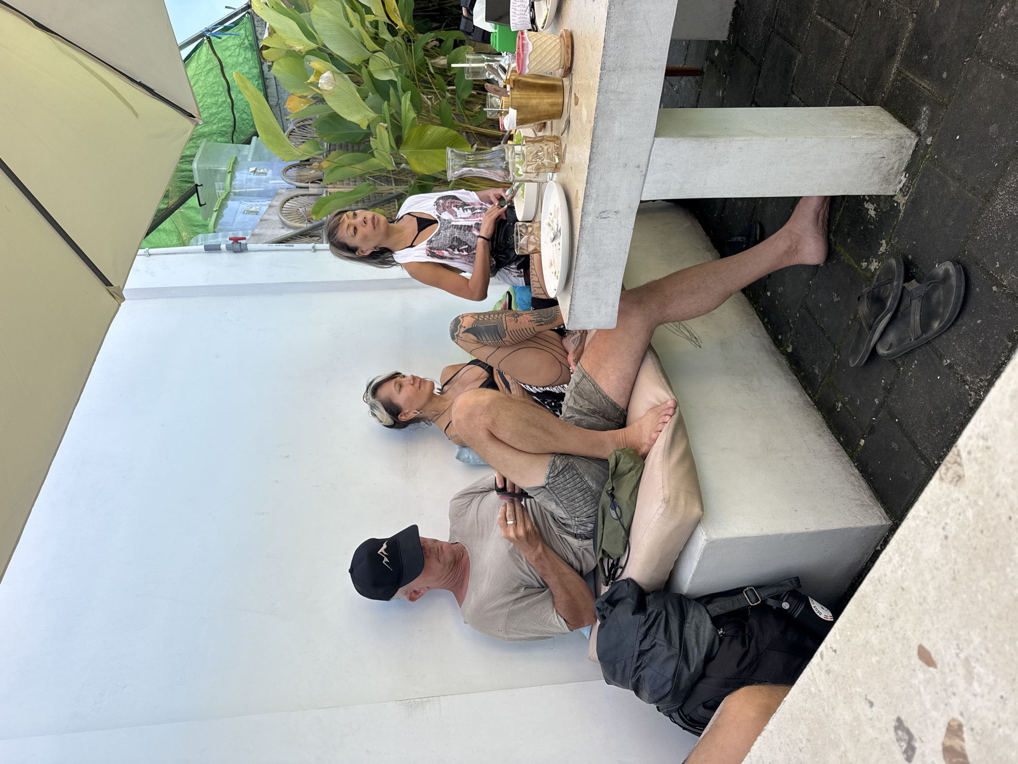 The image shows three individuals seated at an outdoor table in what appears to be a restaurant or cafe in Kuta Selatan, Indonesia. One woman is seated directly at the table, dressed in a white top with a graphic design, and is looking at something in her hands. Beside her, a man with a tattooed leg is seated on the bench, stretching his legs out. Below them, another person, wearing a cap and a light-colored shirt, is looking at a mobile phone. The table contains multiple glasses, plates with leftover food, and decorative containers. The background includes tropical foliage and glimpses of other tables and chairs. The setting appears to be an open-air dining area adjacent to a white wall.