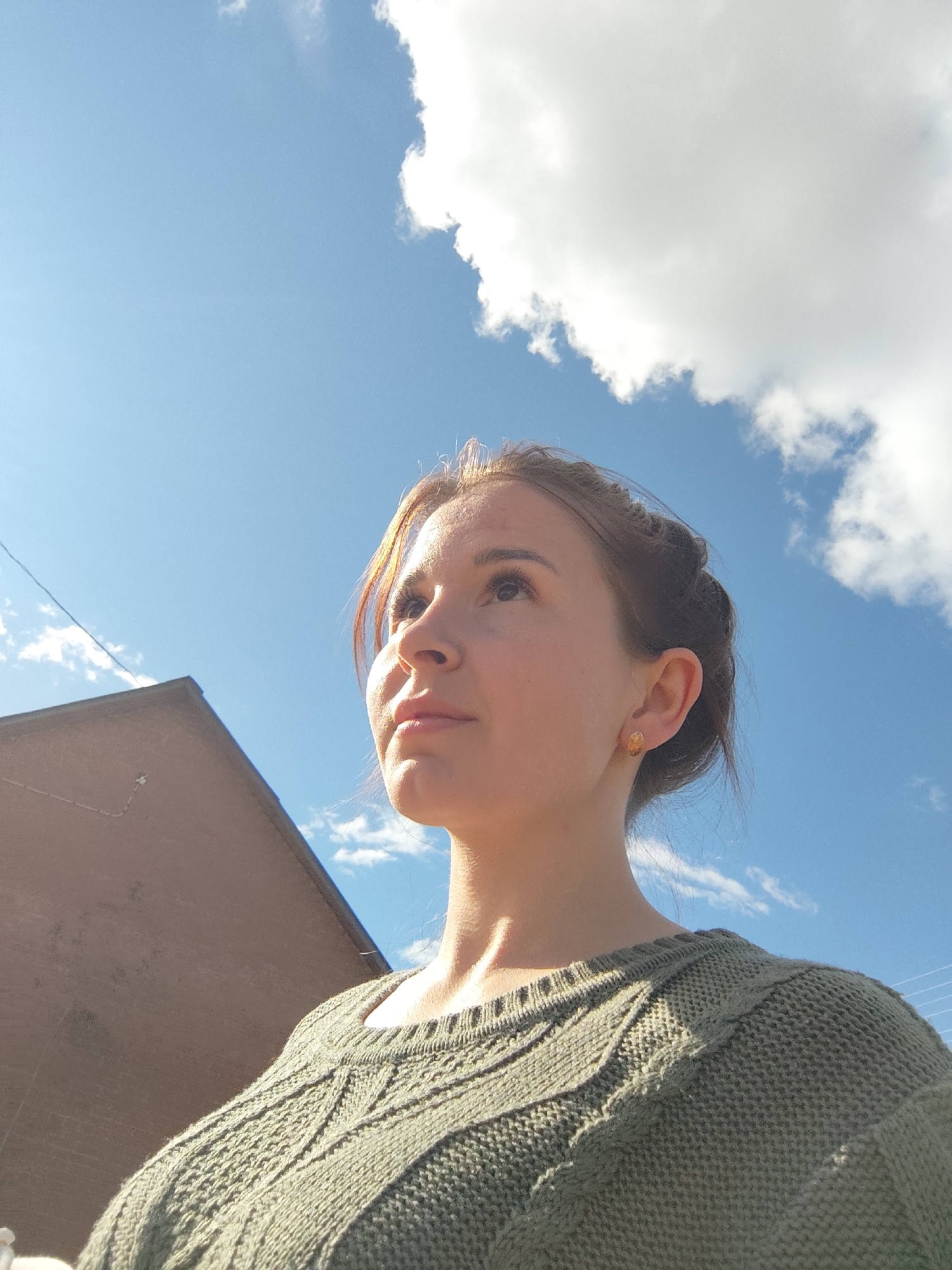 A woman stands outdoors under a partly cloudy blue sky. She is looking upwards. She is wearing a green knit sweater and has earrings. The corner of a red brick building is visible in the lower left of the frame. There are power lines crossing the sky in the background. No indication this image is in Фоки, Russia.