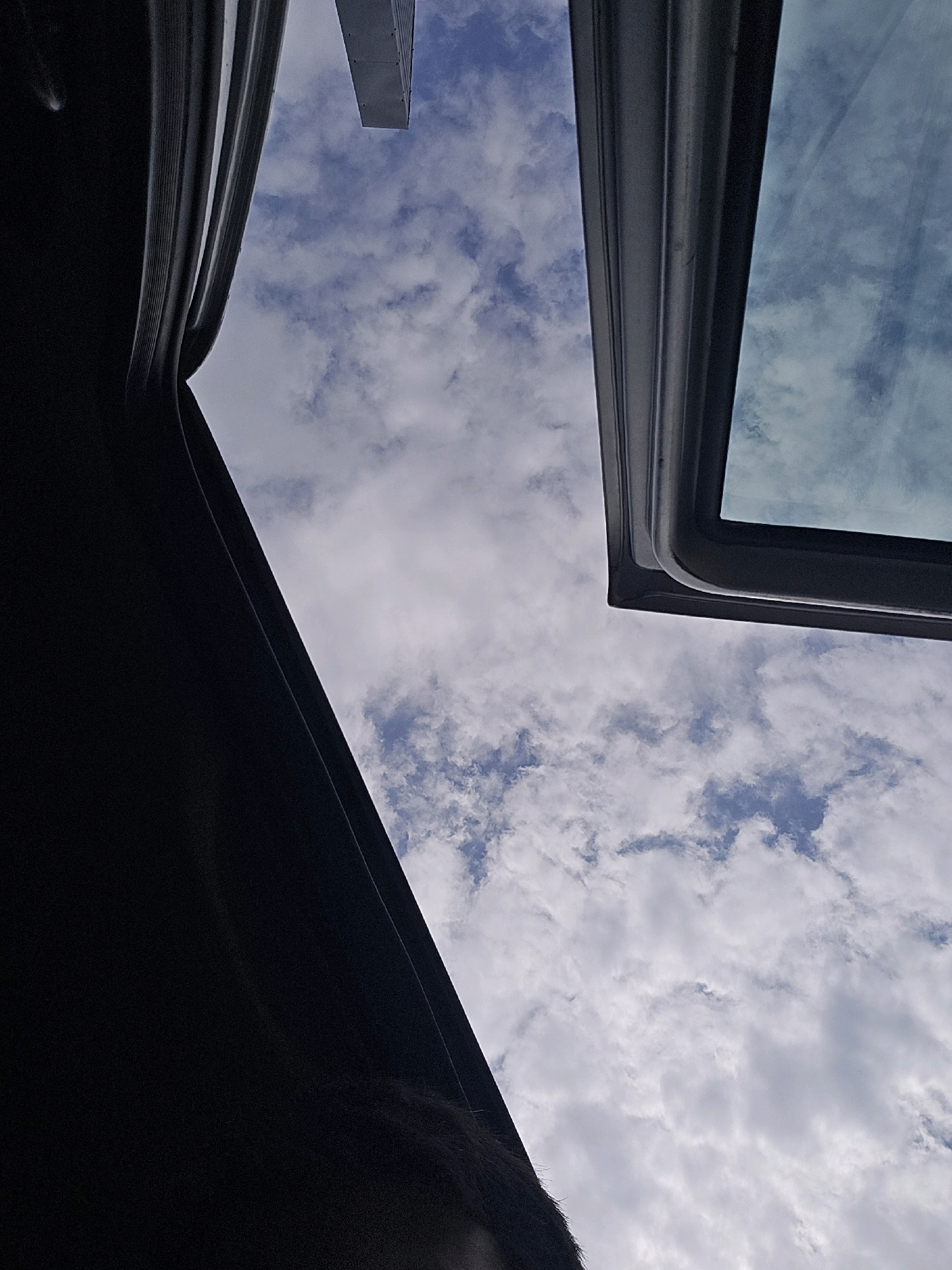 The image is a low-angle shot looking up at the sky from within a vehicle, likely a car, with the sunroof open. The sky is mostly filled with scattered white clouds against a blue background. In the foreground, on the left side of the image, is a dark, curved surface, possibly part of the car's interior trim or a rolled-up shade. On the right side, the edge of the open sunroof frame is visible, and through the glass, more of the sky and clouds are reflected. A sliver of a tall, light-colored building is visible in the upper center of the frame, poking above the sunroof. The scene appears to be set in the city of Avtury, Russia, given the context.