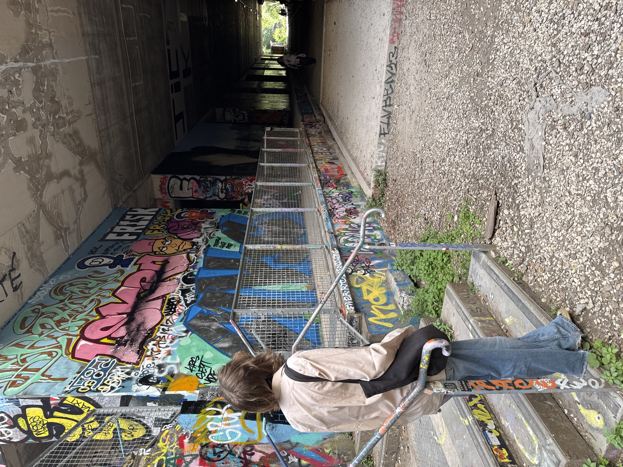 A low-angle, eye-level photograph depicts an individual resting on steps leading into a graffiti-covered underpass or tunnel. The person, seen from behind, has shoulder-length brown hair and is wearing blue jeans, a light tan jacket, and a black bag slung across their shoulder. They appear to be reclining or sitting on the steps, which descend into the tunnel. A metal railing and wire mesh enclosure flank the steps.
The walls of the underpass are heavily covered in graffiti, with a mix of colors and styles of writing. On the right side of the image, beyond the steps and graffiti, is a surface of gravel or small stones. The tunnel extends into the distance, becoming darker and less detailed as it recedes. At the far end, a light source suggests an exit or opening. The photo's perspective is slightly tilted, with the top of the image appearing to lean to the left.
Given the distinctive graffiti styles and the architectural characteristics, the location is likely near Canal Saint-Martin in Paris, France, known for its urban art scene and canal-side underpasses.