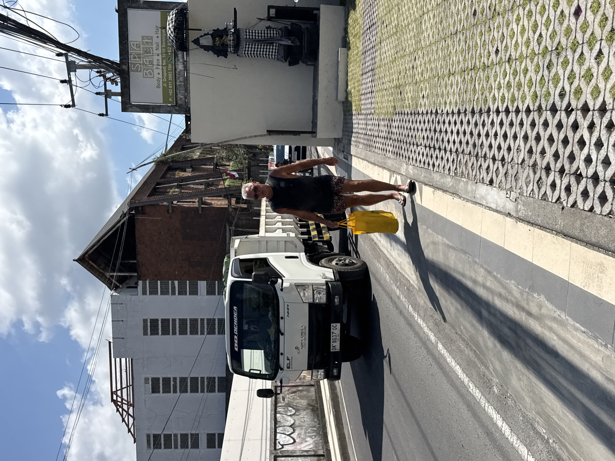An outdoor street scene in what appears to be Kuta, Indonesia, shows a man walking on a sidewalk beside a parked white truck under a bright, partly cloudy sky. The man, of light complexion, wears a black t-shirt, patterned shorts, sunglasses, and flip-flops, carrying a bright yellow shopping bag. Long shadows stretch across the asphalt road and sidewalk, suggesting either morning or late afternoon.
The white Isuzu Elf truck, visible on the left side of the frame, bears the text "DO DENDEA," "ISUZU," "ELF," and "NMR," along with a license plate "DK 8017 OC" and a "05-30" marking.
Behind the man, a white building wall features a traditional Balinese statue or shrine adorned with a black and white checkered cloth. Above the shrine, a rectangular sign for "SPA BAEL" advertises "Body • Face • Nail • Hair" services, listing an address as "Jl. Drupadi No. 1010 Seminyak" and phone numbers "+62 811 3993 701" and "+62 85772." To the right of the sidewalk, a textured concrete grid with grass growing through its openings forms a ground cover.
In the background, tangled utility wires crisscross against the sky, connected to poles, and a multi-story building with small windows and a dark, gabled roof structure is visible. The overall setting captures a typical urban street in Bali.