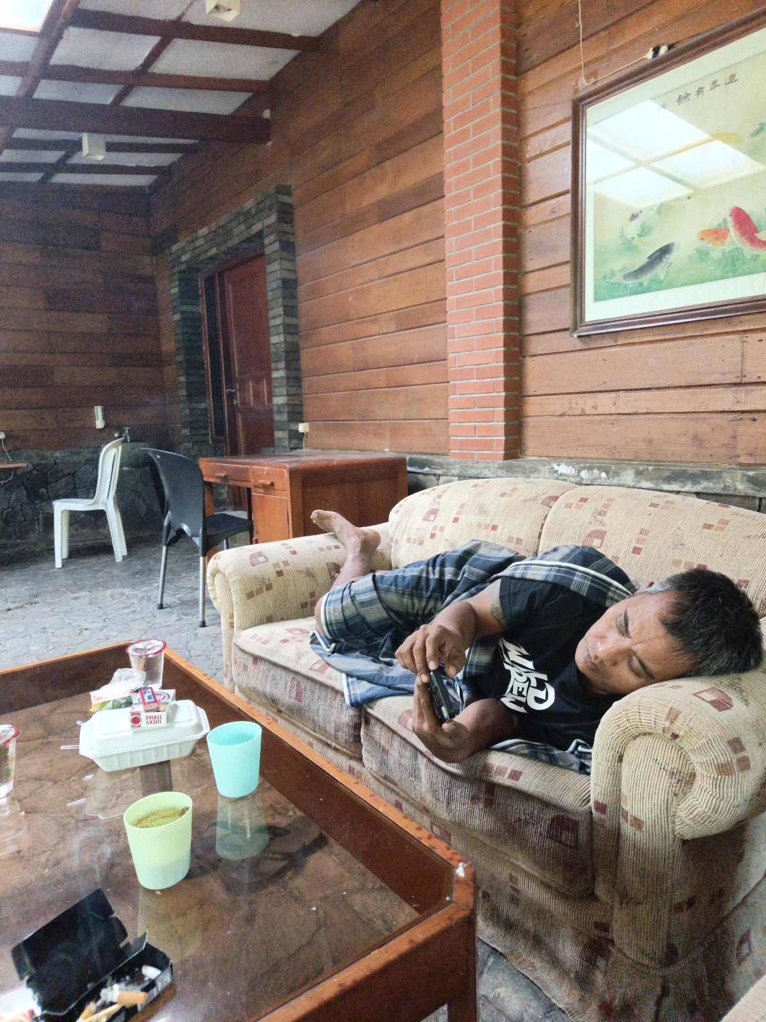 A man is lying on his side on a patterned sofa, holding a smartphone in his right hand. He is wearing a black t-shirt with white lettering and plaid shorts. His eyes are closed, and he appears to be resting or sleeping. The sofa is located outdoors on a patio area with wooden walls and a tiled floor. A wooden coffee table is in front of the sofa, holding two plastic cups, a white takeout container, and an open black container with cigarette butts. In the background, there is a wooden door, two chairs, and a framed painting of fish on the wall. The overall setting suggests a casual, relaxed environment, possibly a vacation home or a resort in Lembang, Indonesia.