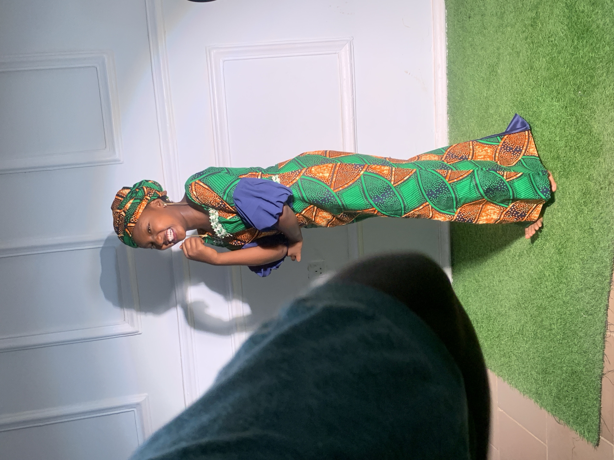 The image shows a young woman smiling and wearing a traditional African dress with a matching headwrap. The dress has a vibrant green and orange pattern with blue sleeves. She appears to be standing indoors against a white wall with decorative molding. The floor is partially covered with green artificial turf. A portion of a person's dark green clothing is visible in the foreground, suggesting the photo was taken at close range. This photograph was taken in Barakin Akawo, Nigeria.
