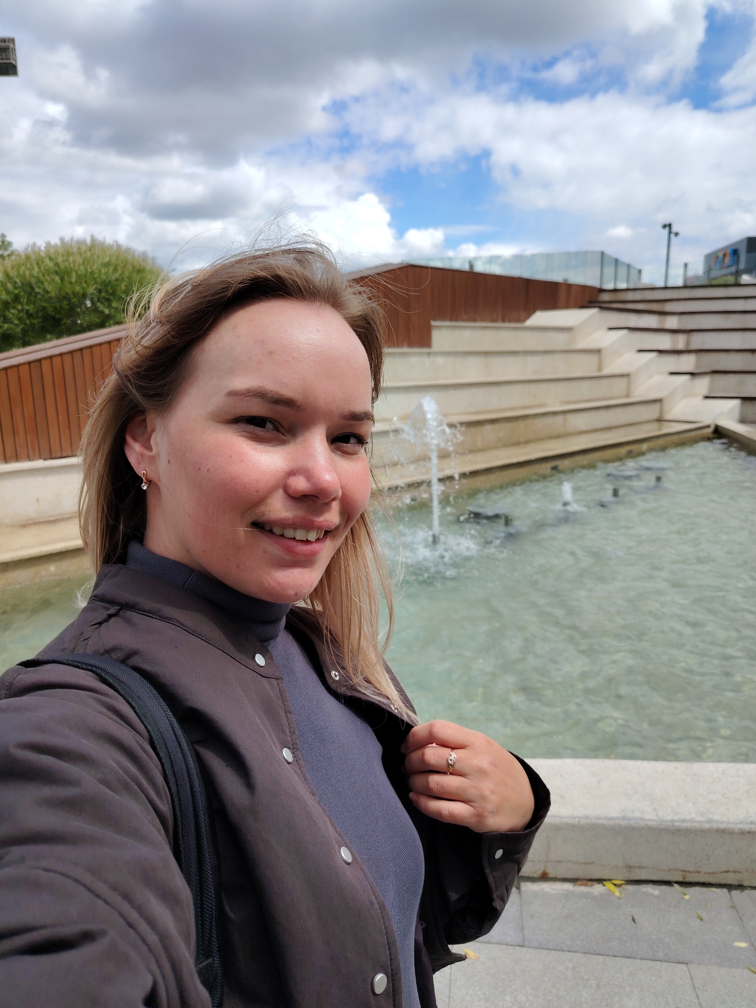 The image shows a young woman in a selfie near a fountain. She has fair skin, blonde hair, and is wearing a dark brown jacket over a gray turtleneck. She has a ring on her right hand and earrings. The fountain is tiered with water features and is set against a backdrop of modern architecture and a cloudy sky. The location is likely in Yekaterinburg, Russia.