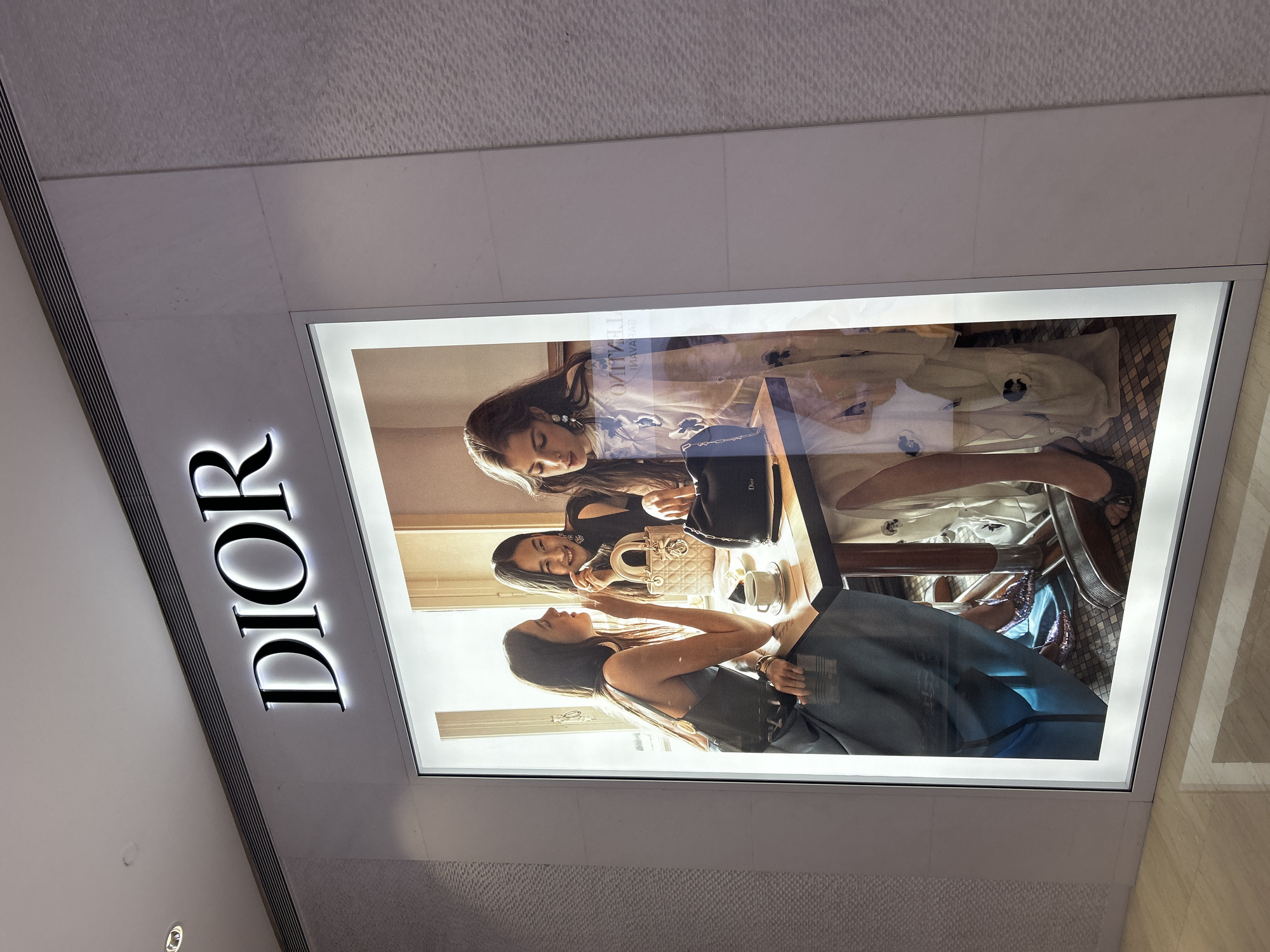 The image shows a Dior store sign mounted on a wall, likely in a shopping mall. The Dior logo is illuminated and placed next to a rectangular advertisement display showing three women sitting at a table. There are purses, cups, and other objects on the table. The location is probably Kuala Lumpur, Malaysia, although the surrounding environment is not visible in detail to verify this.