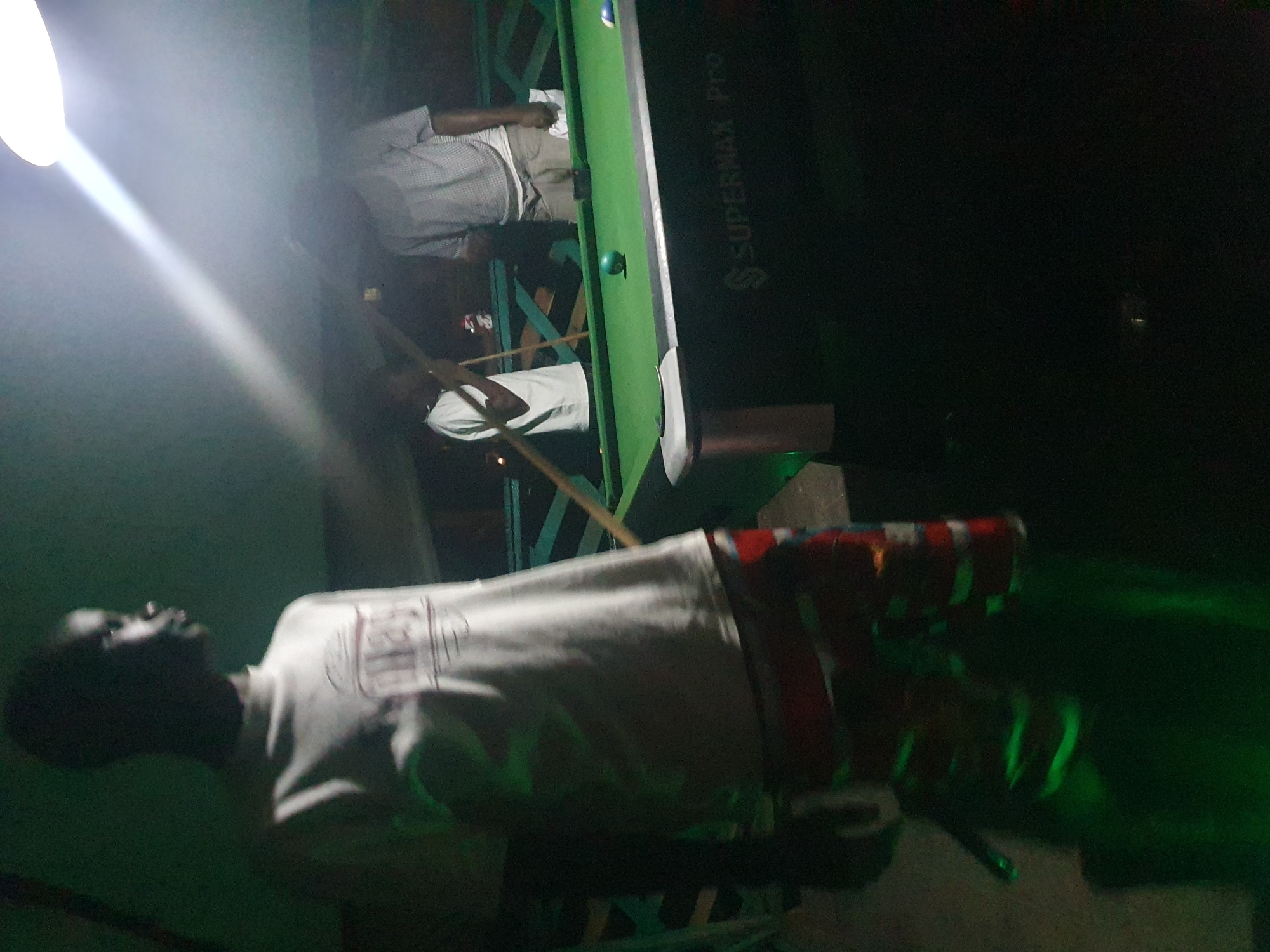 A group of people are playing billiards on a green pool table under a bright overhead light. The table is labeled "SUPERMAX Pro". A young man in the foreground wears a white t-shirt and colorful shorts. Two other men are visible in the background, one with a pool cue. The environment appears to be an outdoor setting, possibly a bar or recreational area, with some greenery and wooden structures visible. The scene is dimly lit, with the overhead light casting strong shadows. The image quality is grainy and slightly out of focus.