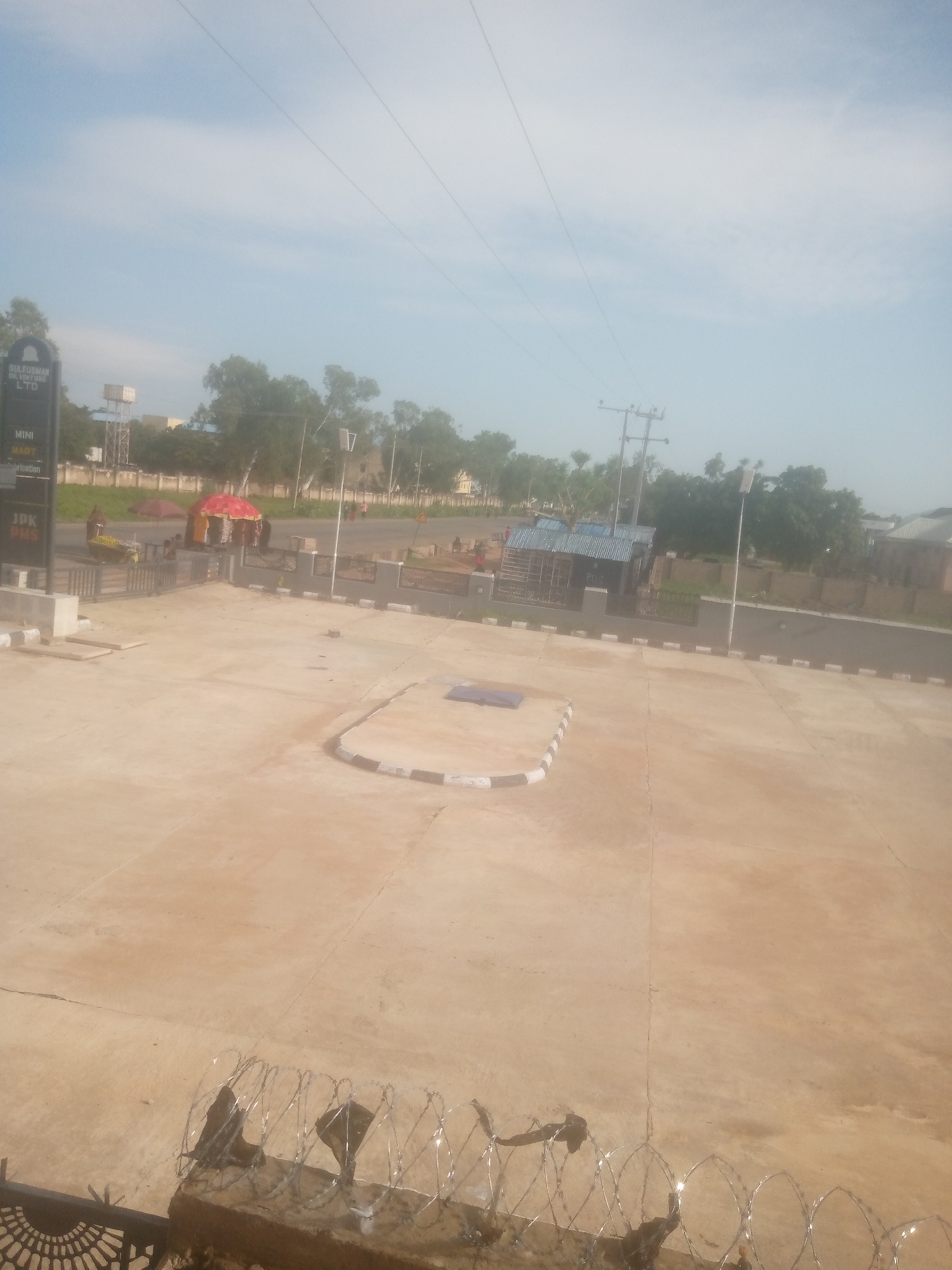 The image shows an outdoor scene in Jalingo, Nigeria. A paved area in the foreground, possibly a parking lot or plaza, is bordered by barbed wire and a low decorative fence. In the mid-ground, a street runs alongside a gray wall with a checkered border. Several buildings are visible, including one with a blue metal roof. A tall utility pole with wires stands near the street. A red umbrella and a yellow awning are set up in an open area, with several people gathered around them. To the left, a black signpost with white text indicates "SULEIMAN OIL VENTURE LTD," "MINI MART," and "JPK HMS." Trees and a water tower are present in the background, under a lightly clouded sky.