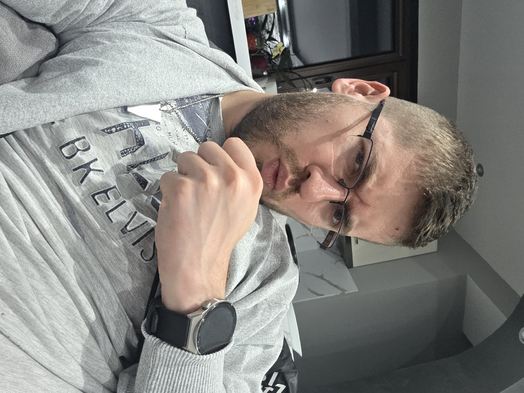 An indoor, close-up shot captures a man in Yagodovo, Bulgaria, looking directly at the camera with a neutral and somewhat pensive expression. The man has short, light brown hair and a trimmed beard. He wears black-framed, rectangular glasses. His right hand is raised to his chin or mouth area, with his fingers lightly touching his face, suggesting contemplation.
He is dressed in a light grey hooded sweatshirt worn over a lighter grey t-shirt. A silver chain is visible around his neck, peeking out from under his t-shirt. The front of his t-shirt displays partial text that reads "BK ELVIS". On his left wrist, he wears a black smartwatch featuring a round, dark screen, a metallic silver bezel, and a black strap.
The background is indistinct but appears to be a modern indoor space. Elements such as a dark window frame, light grey walls, and subtle reflections suggest artificial indoor lighting. The ambient light is soft, contributing to the composed mood of the portrait. The image was originally captured with a sideways orientation.