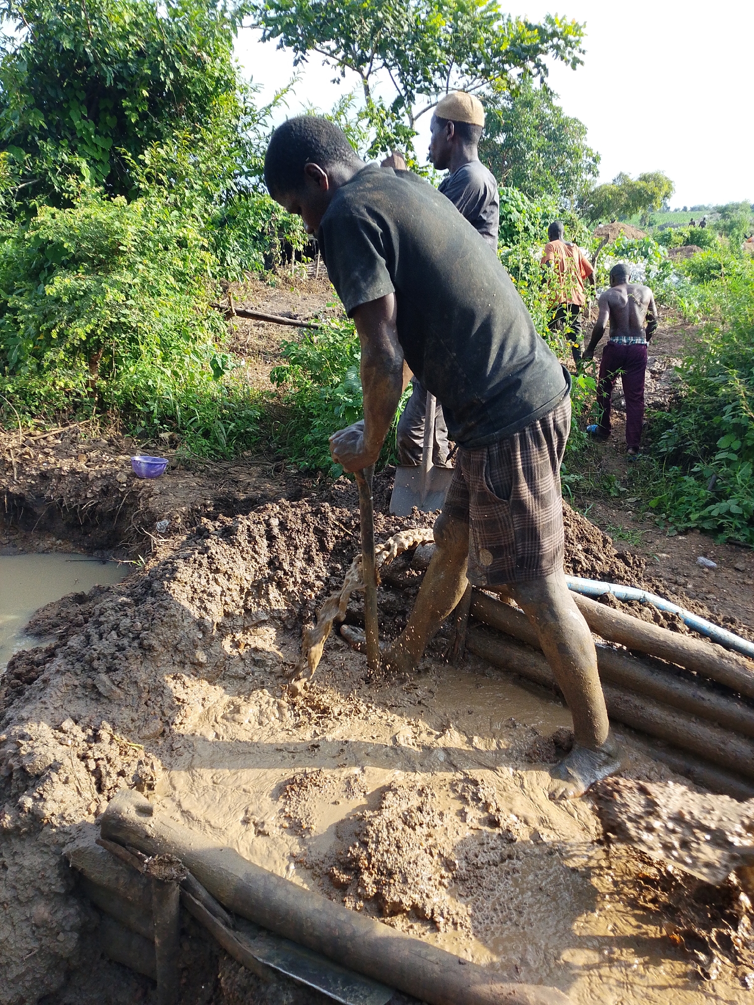 In Otulu, Nigeria, amidst lush green vegetation and hilly terrain, several individuals are engaged in labor, likely related to excavation or construction. The central figure, a Black male wearing a dark t-shirt and plaid shorts, is actively digging in a muddy pit with a shovel. His legs and lower body are covered in mud, indicating strenuous work. The pit itself is filled with murky water and thick mud, with wooden logs and pipes surrounding it, suggesting some form of infrastructure development or water management. In the background, other men are visible, some also appearing to be working in similar conditions, while others observe the activity. The overall scene depicts a rural, outdoor environment under natural sunlight, with a focus on manual labor and the challenges of working with the earth in a tropical setting.