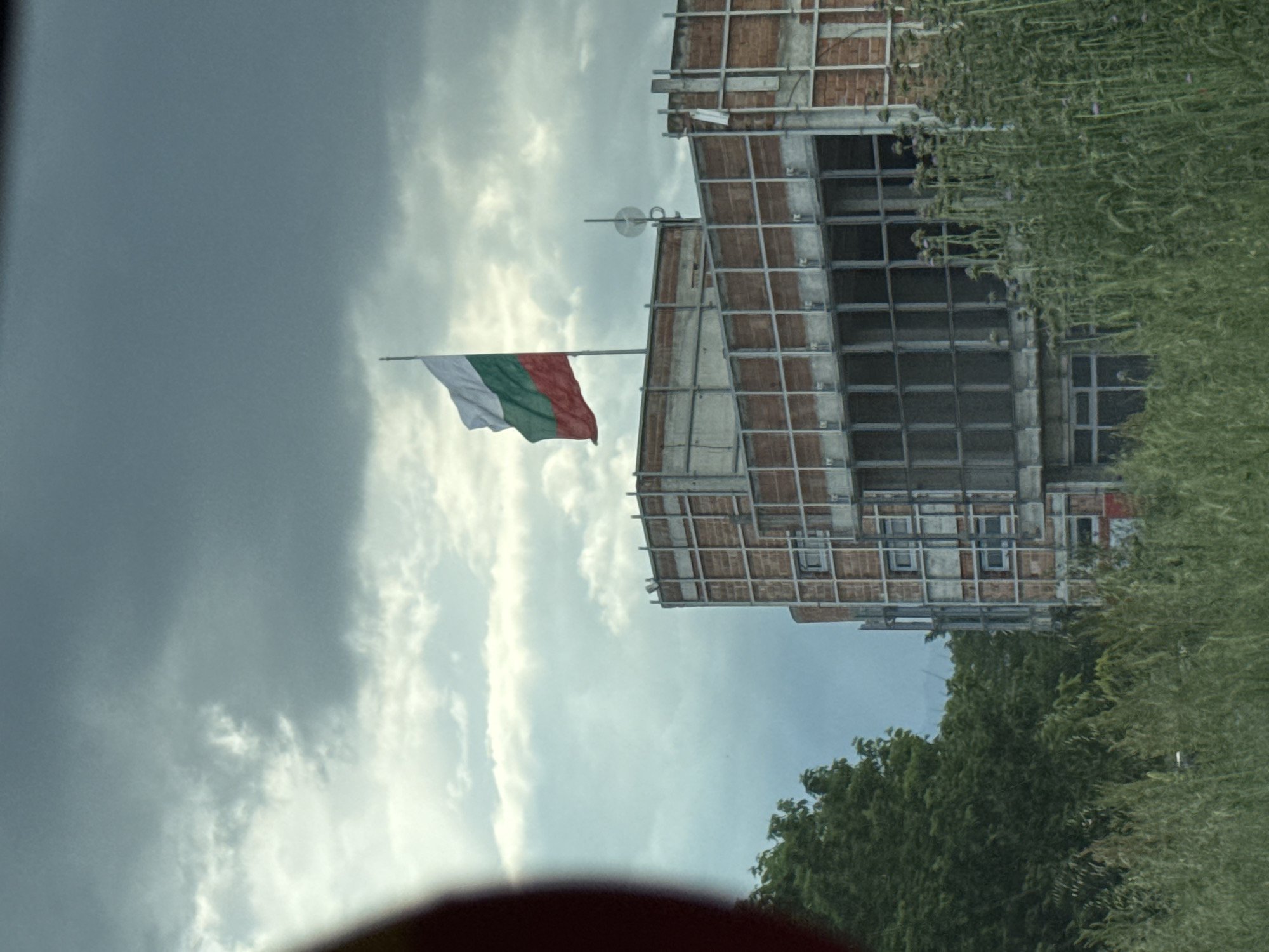 The image shows a building with a steel frame under construction, likely in Пловдив, Bulgaria, as the Bulgarian flag is hoisted on a pole attached to the building's structure. Overgrown vegetation is visible to the right, suggesting the building is on the outskirts of a developed area. The sky is overcast with a mix of dark and light clouds. A red object is partially visible in the foreground, possibly part of a vehicle's interior or exterior.