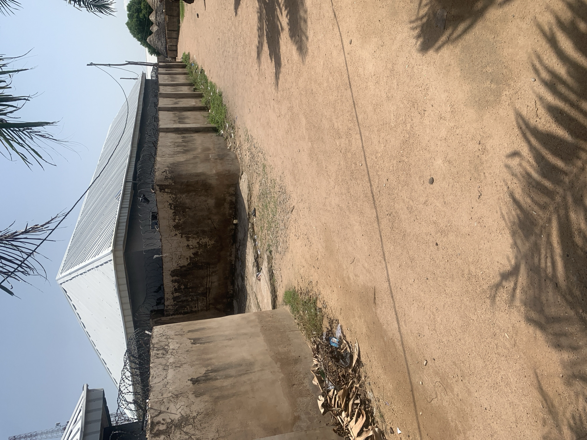 A dirt road winds through a community in Jalingo, Nigeria, bordered on the left by a tall, weathered concrete wall topped with coiled barbed wire. Behind this wall, a modern building with a corrugated metal roof is visible. To the right of the road, thatched-roof huts create a traditional village scene. The ground is dry and sandy, with scattered debris, including dried leaves and plastic waste, near the base of the concrete wall. Palm fronds cast shadows on the road. A utility pole with wires stands along the right side of the road. The sky is clear and bright blue.