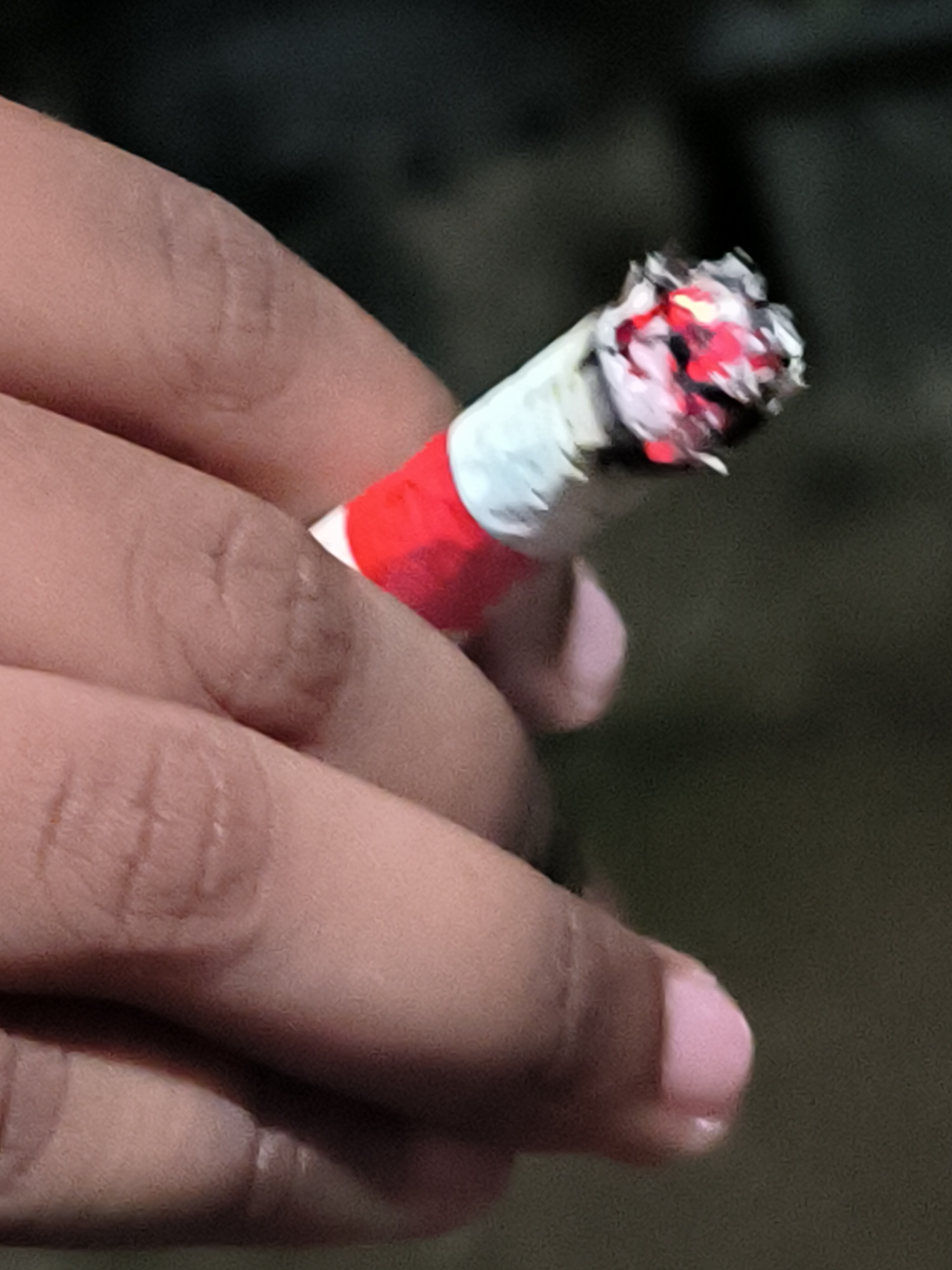 The image shows a close-up of a hand holding a lit cigarette. The cigarette has a white paper wrapper and a red band near the tip. The tip of the cigarette is glowing red and appears to be burning. The hand is light-skinned, and the fingernails are painted pink. The background is dark and out of focus, suggesting an indoor or nighttime setting. There are no discernible environmental details that would indicate a specific location within Lhokseumawe, Indonesia.