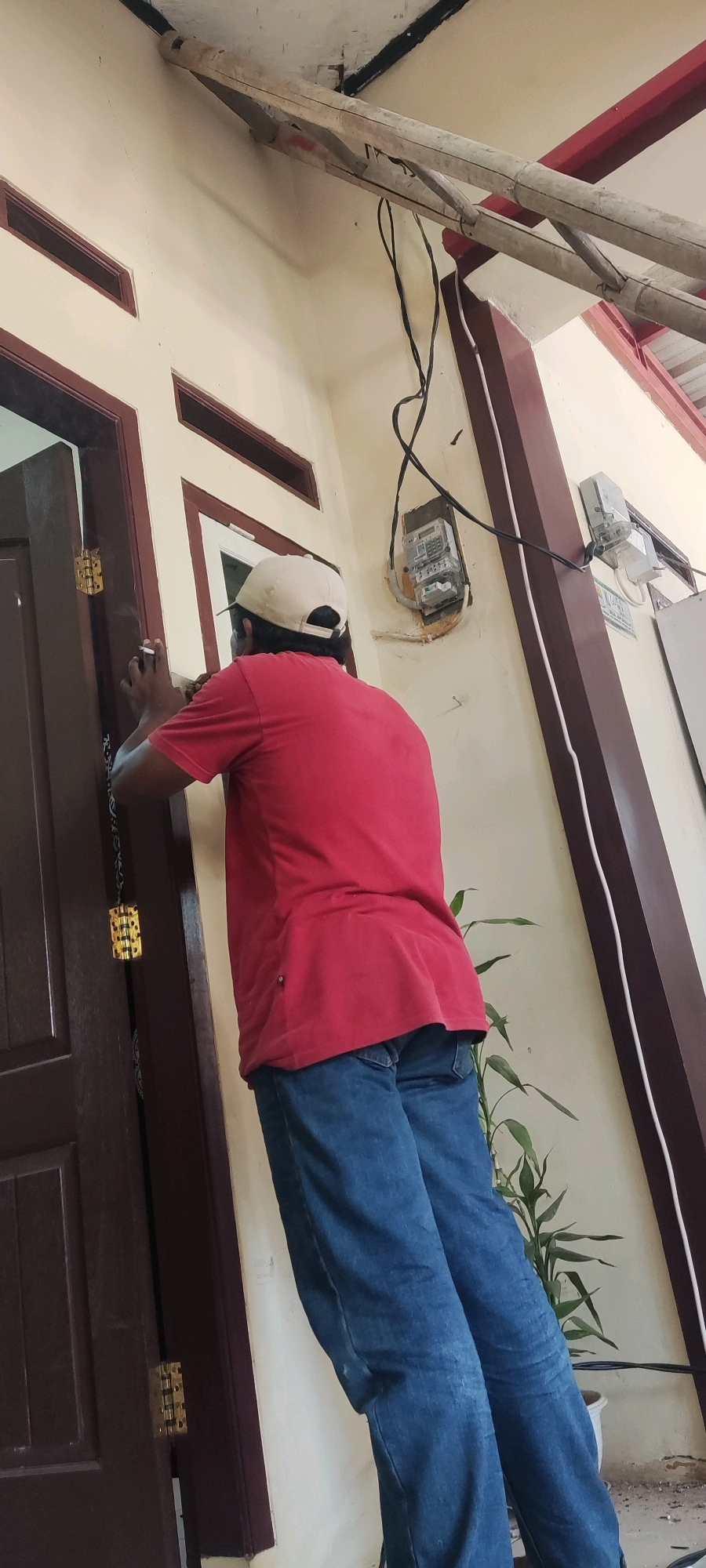 In Japura Kidul, Indonesia, a man wearing a red t-shirt and blue jeans stands facing away from the camera, appearing to be working on a brown doorframe. He is wearing a light-colored baseball cap. His right hand is near the door, and he seems to be holding a cigarette in his left hand, which is raised to his face. Visible on the wall behind him are two electricity meters. One meter, older in design, is mounted on a brown backing, with several black wires connected to it. Another, more modern-looking meter is mounted to the right. A tall green plant in a white pot is situated near the right side of the frame. The building's exterior features a light-colored wall with dark brown trim around the windows and door. Overhead, a wooden beam construction is visible, with black electrical wires running across it.