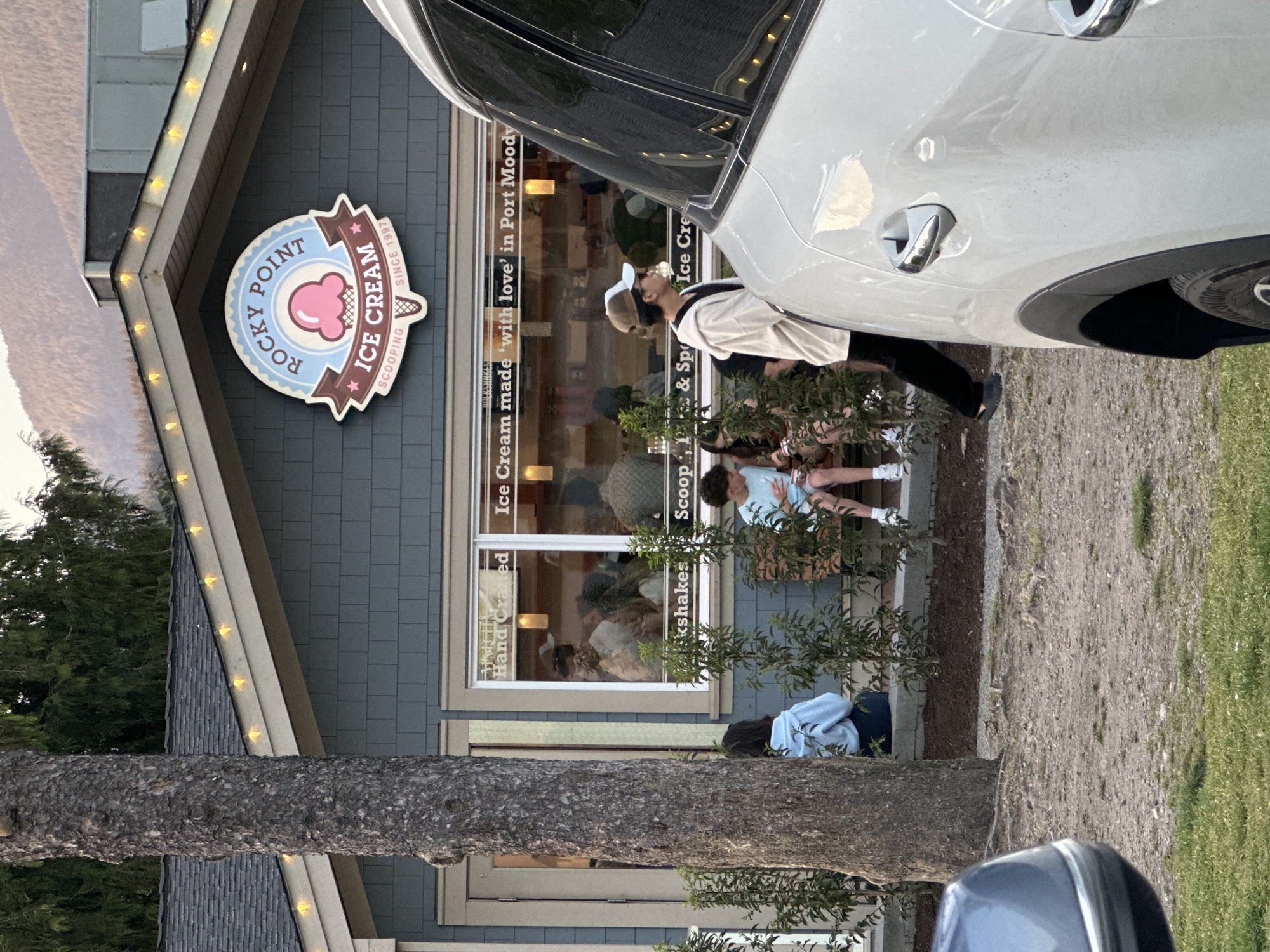 The image shows the Rocky Point Ice Cream shop in Port Moody, Canada. The shop's blue and pink sign is visible. A light-colored car is partially obstructing the view of the shop's storefront window, which displays the words "Ice Cream made 'with love' in Port Moody", "Scoops and Shakes", "Ice Cream", and "Hand Crafted". Several people are seated outside the shop, including a person wearing a beige hat and a white top. A tree is positioned in front of the building, obscuring part of the lower section of the storefront and a parked car in the bottom foreground. The environment appears to be in a semi-urban setting with trees, shrubbery and a road nearby.