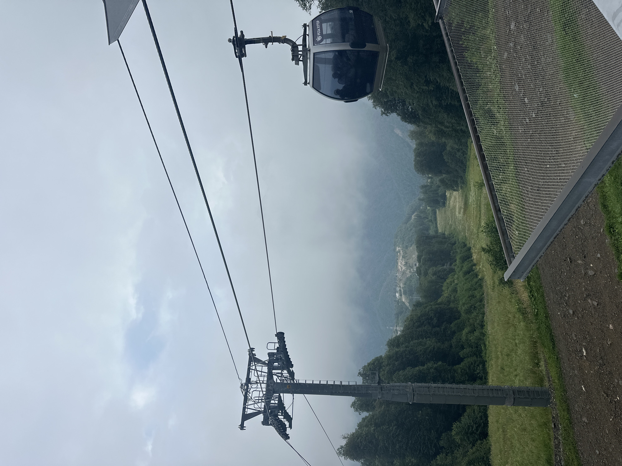 The image captures a scene in Estosadok, Russia, featuring a cable car ascending a mountainous terrain. The cable car, a modern gondola with tinted windows, is suspended from a thick cable, with a supporting pylon visible on the left. The gondola has "РОЗА ХУТОР" written on its side, indicating its location at the Rosa Khutor resort. Inside the gondola, silhouettes of passengers can be seen. The surrounding environment is characterized by lush green slopes covered with dense forestation and a valley below that appears to be a ski slope in warmer months, given the manicured appearance of the grassy area. The sky is overcast, with a hazy, diffused light suggesting a cloudy or foggy day, which partially obscures the distant mountain ranges. In the foreground, there is a metallic railing with a mesh netting, possibly part of a viewing platform or safety barrier for a hiking or observation area. The overall impression is one of a scenic, natural landscape enhanced by modern recreational infrastructure.