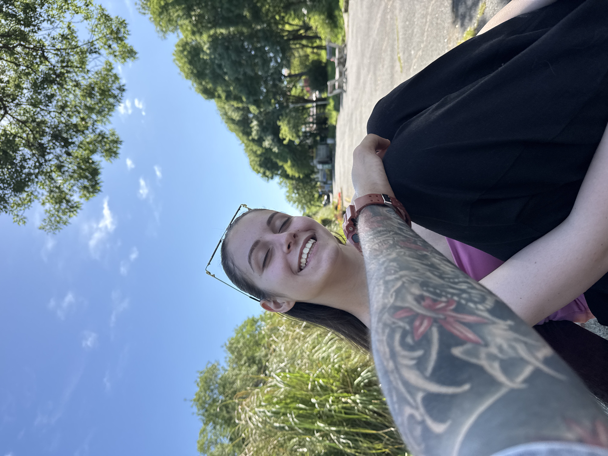 A low-angle outdoor shot shows a smiling woman with a full tattoo sleeve on her left arm. The scene appears to be taken in a park or outdoor area in daylight, possibly in Tsarevo, Bulgaria. The background features trees, sky, and a distant glimpse of park benches. The woman is wearing eyeglasses and has long hair. She is seated, as a portion of black clothing is visible. Her sleeve has a detailed floral tattoo. The sky is blue with few clouds, suggesting a sunny day.