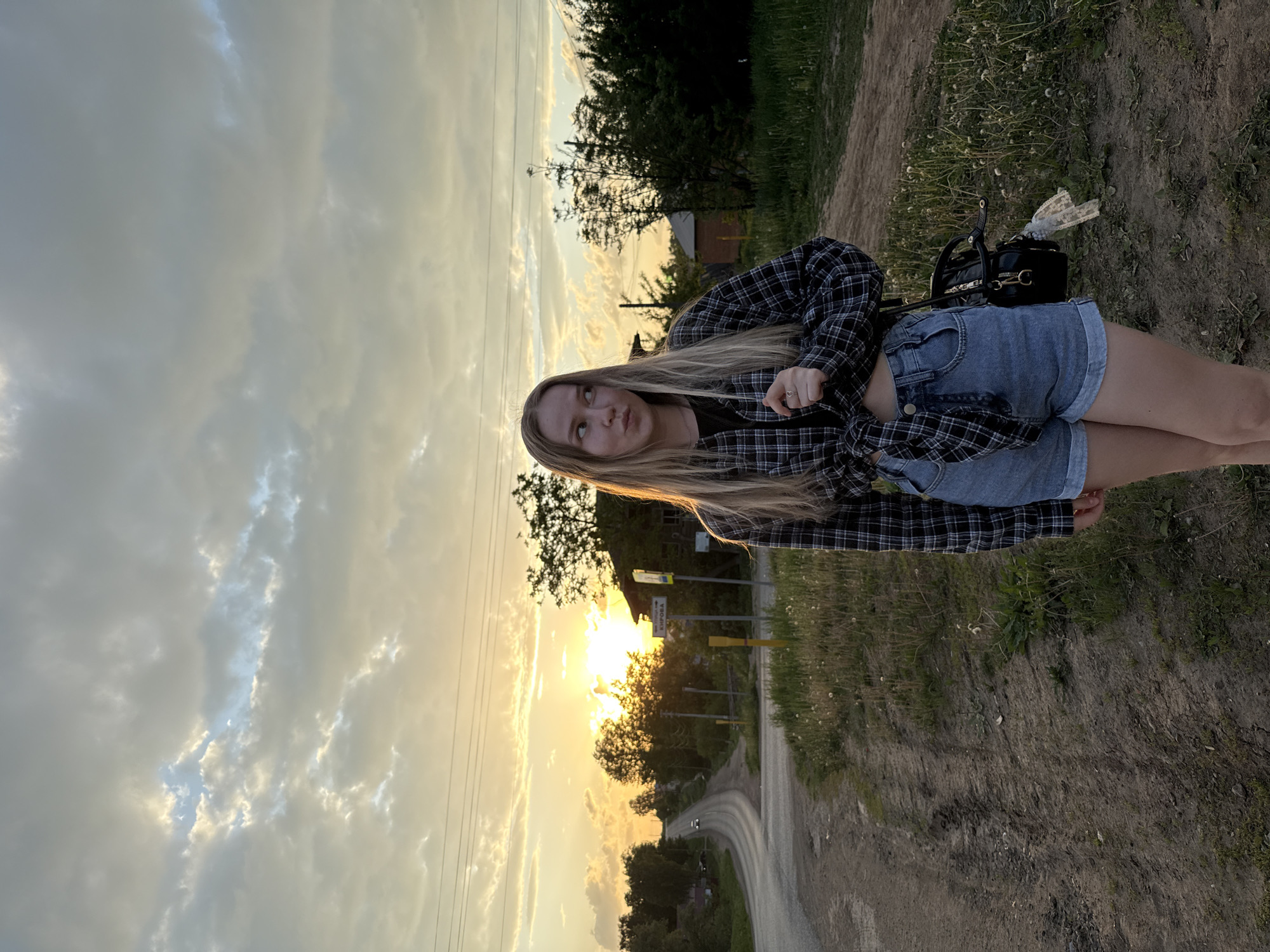 A young woman with long blonde hair stands on a dirt road at sunset. She wears a black and white plaid shirt over a black top and denim shorts. A black purse sits on the ground near her. In the background is a road, trees, and the sun setting, with a sign that reads "КИРОВА". Power lines are visible in the sky. Based on the road sign, the location is in the city of Фоки, Russia.