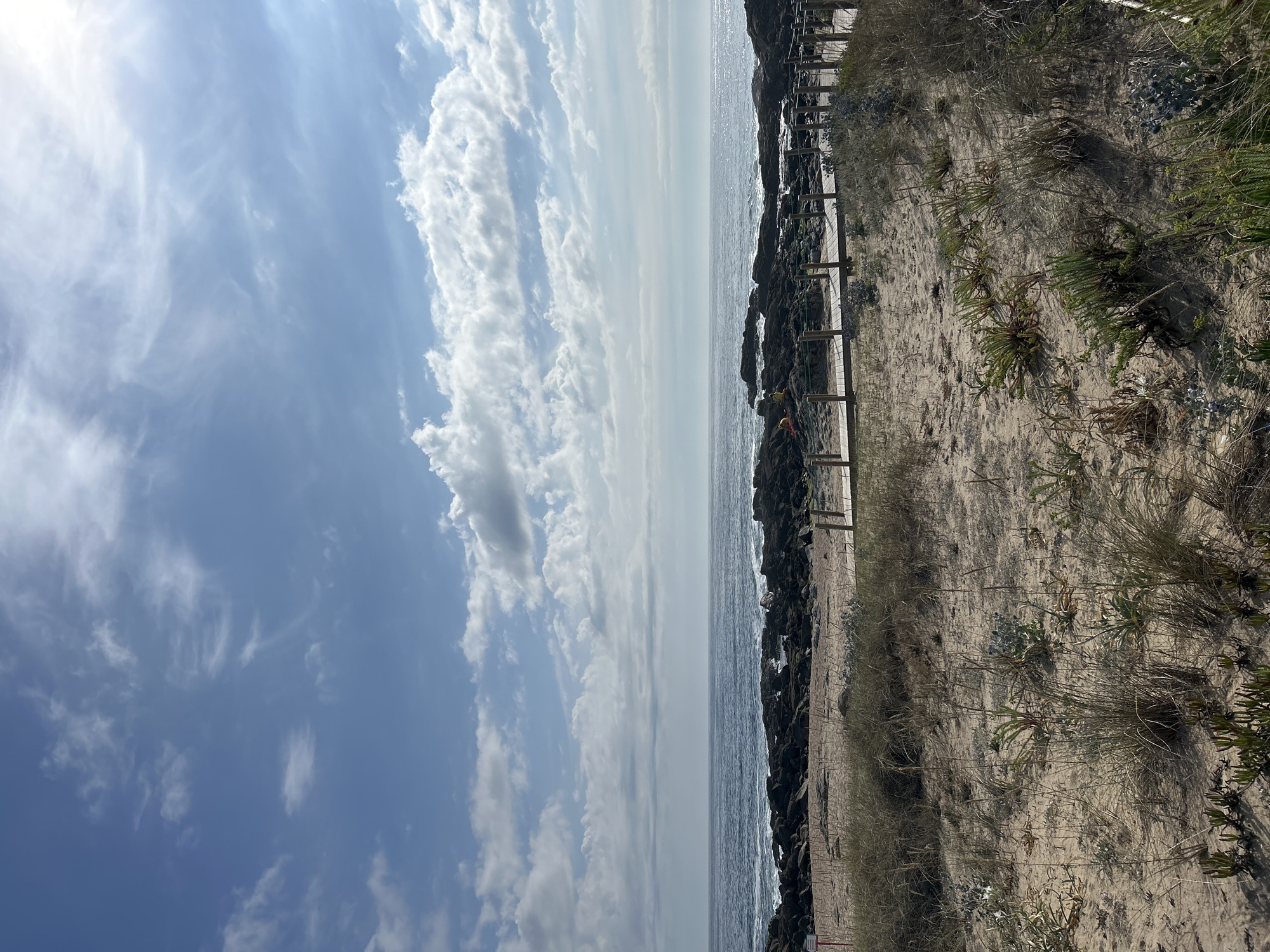 A scenic view of a beach with sand dunes, a wooden boardwalk, rocky shore, and the ocean under a partly cloudy sky. The sand in the foreground is covered with sparse, dry grass and some green vegetation. A wooden boardwalk with a railing runs diagonally from the lower right corner towards the left. Beyond the sand dunes and boardwalk, a rocky shoreline meets the ocean. The ocean appears calm with small waves. The sky is blue with scattered white clouds, some of which are illuminated by sunlight. In the distance, on the rocks, two figures in yellow are visible, possibly lifeguards or beachgoers.