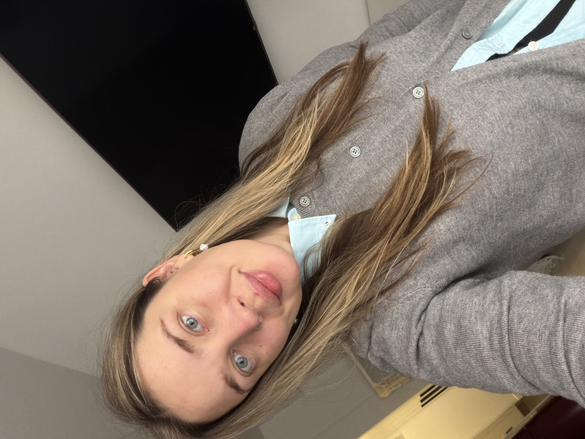 A woman with light blonde hair and blue eyes is shown in a close-up, angled portrait. She is wearing a light blue collared shirt under a gray cardigan. The background consists of a black rectangle, likely a television screen, and a portion of a beige-colored object, possibly furniture or a wall. No city or Russian context is evident in the frame.