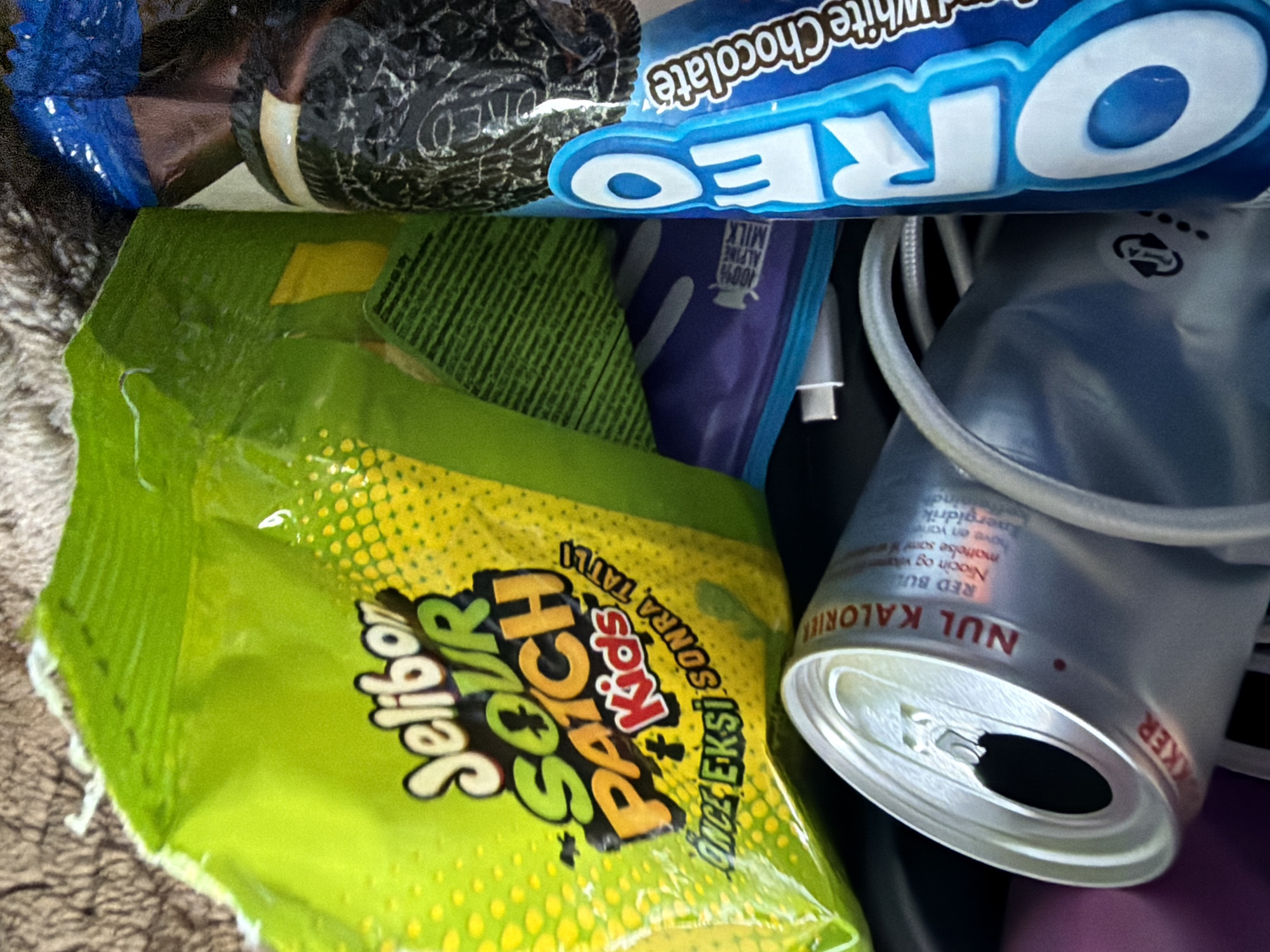 The image shows a close-up of several snack items, including a bag of Sour Patch Kids candies, a package of Oreo cookies, and an opened can of Red Bull. The items are arranged on a textured surface, possibly a carpet or fabric. The Sour Patch Kids bag is green and yellow, with the brand name prominently displayed. The Oreo package is dark blue and white, with the Oreo logo visible. The Red Bull can is silver and red, with the brand name and some text in a foreign language. The lighting is dim, and the focus is sharp on the snack items. There are no people or clear indications of a specific location, such as a city or environment.