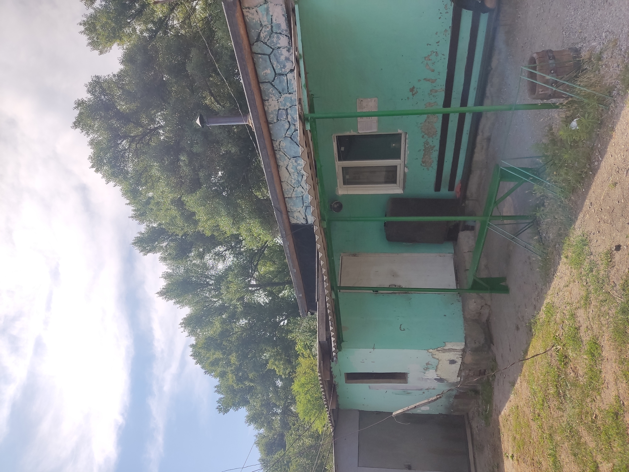 The image depicts the exterior of a small, weathered building painted a light teal color, situated in the village of Radisne, Ukraine. The building appears to be a single-story structure with a corrugated metal roof that extends to form a covered porch area. The walls show signs of wear, with peeling paint revealing older layers and possibly some brickwork underneath.
To the left of the main building is a larger, grey metal garage door. The main building features a white door on the covered porch, and a single, rectangular window with a white frame is visible on the right side of the structure. Above the window, the upper part of the building is decorated with a band of faux stone cladding, painted in shades of blue and grey, which is also showing signs of deterioration. A dark metal chimney extends upwards from the roof.
In front of the building, a green metal bench is positioned under the porch. The ground in front is a mixture of dirt and gravel, with patches of dry grass and weeds. There is a dark wooden barrel visible on the right side of the image, and some green metal supports for what appears to be an awning or additional roof structure are also present. The background is dominated by a dense canopy of green trees, suggesting the location is either a rural area or a park. The sky above is partially cloudy, with bright sunlight indicating daytime. No people are visible in the image.