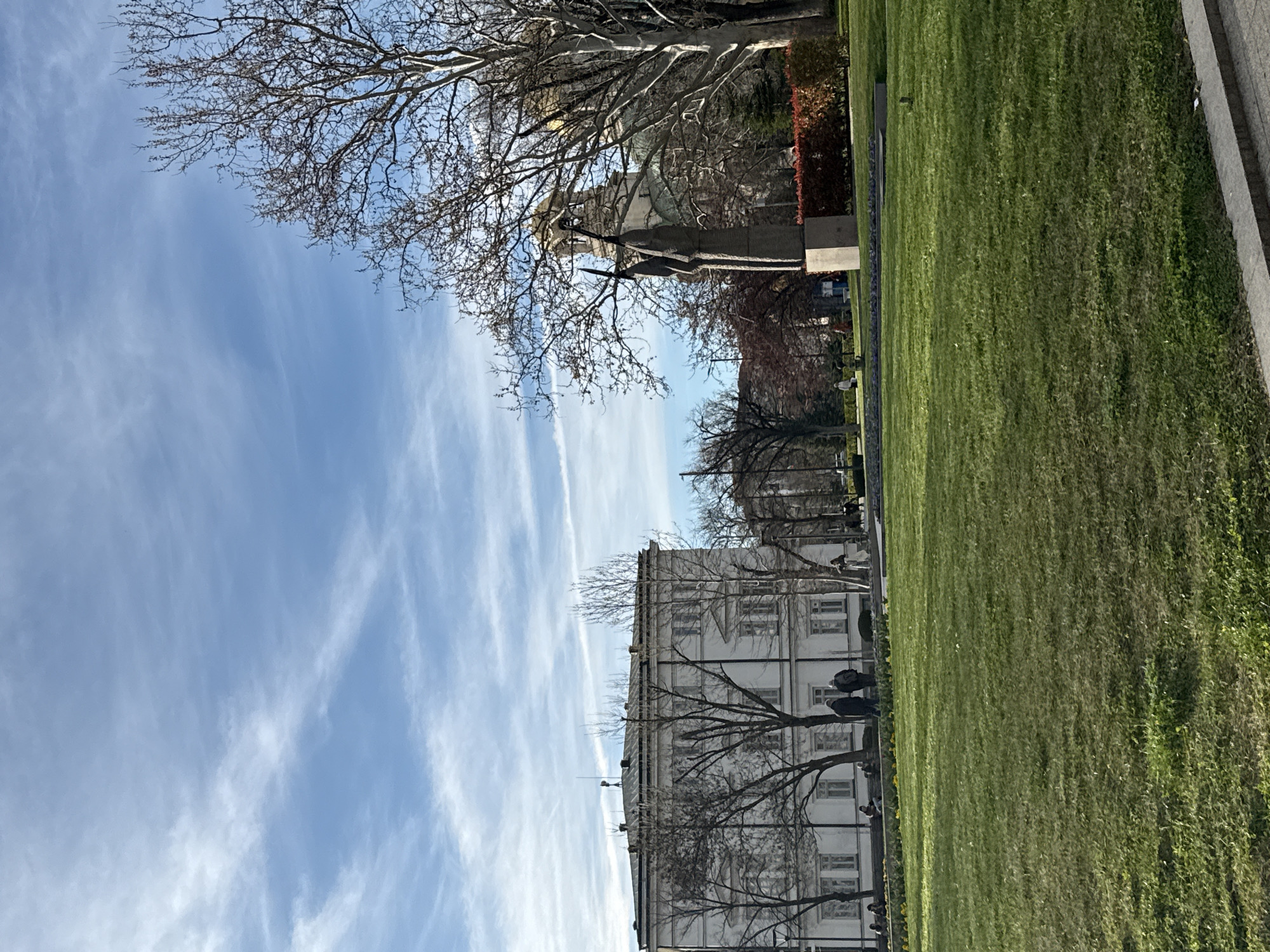 The image is a landscape-style photograph, rotated counterclockwise by 90 degrees. A wide expanse of green grass fills the right side of the frame. A pathway made of rectangular blocks lies along the top edge of the lawn.
To the left of the lawn and path is a large, white, multi-story building with rows of windows. A few bare trees stand in front of the building, their branches obscuring the facade in places. People are visible on the sidewalk near the building. Pigeons are on the grass next to the building.
To the left of the building, the sky is visible. It is a light blue color with wispy, white clouds streaking across it.
In the upper right corner is a statue in a park-like setting with bare trees. A red hedge is visible next to the statue.
The image appears to have been taken in Borisova gradina park in Sofia, Bulgaria. The statue could be the Bulgarian writer and journalist, Hristo Botev. The building is most likely the National Assembly of Bulgaria.