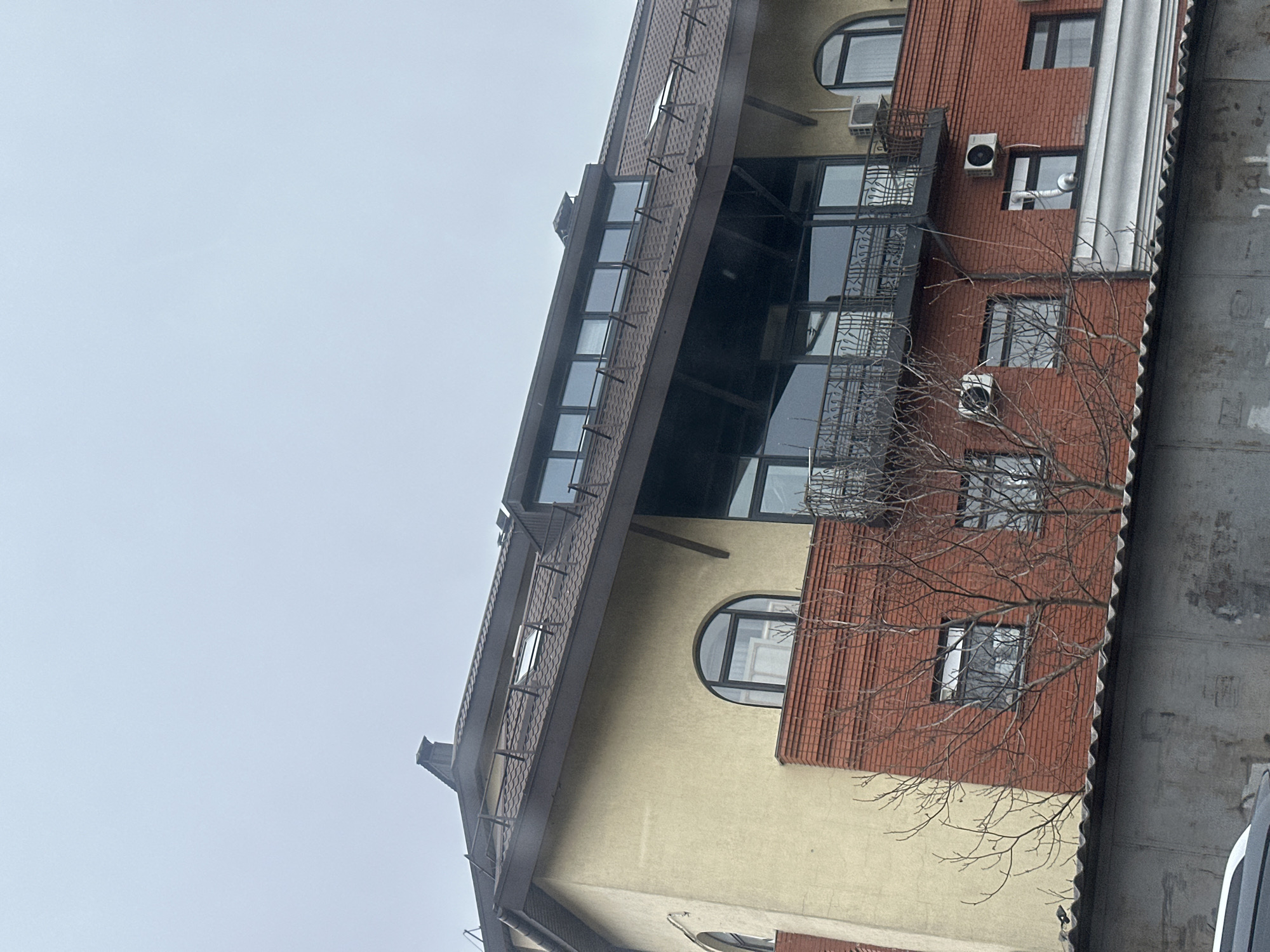 The image shows a multi-story building with a pitched roof and a combination of beige stucco and red brick facade. The building features a series of windows, including arched ones, and balconies with metal railings. Air conditioning units are visible on the exterior walls. A bare tree branch is in the foreground, partially obscuring the building. The sky is overcast. The building's architectural style and the presence of Ukrainian text may indicate it is located in Kyiv, Ukraine. A section of the building is partially hidden with light gray and white elements, perhaps an adjacent structure or vehicle.