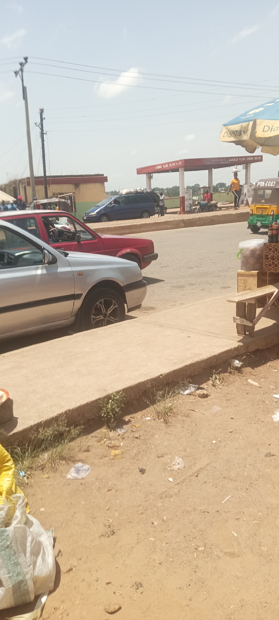 The image depicts a street scene in Kuru Dungus, Nigeria. A silver Volkswagen Golf and a red Volkswagen Golf are parked along the side of the road. Across the street is a petrol station with people getting petrol on a motorbike and filling their car with gas. A blue minivan also has items on top of it. A yellow auto rickshaw is also parked nearby. On the right side of the image, a wooden table sits next to the sidewalk, with a blue and yellow sun umbrella providing shade. The ground is sandy and littered with trash. The sky is partially cloudy. A power line runs along the road.