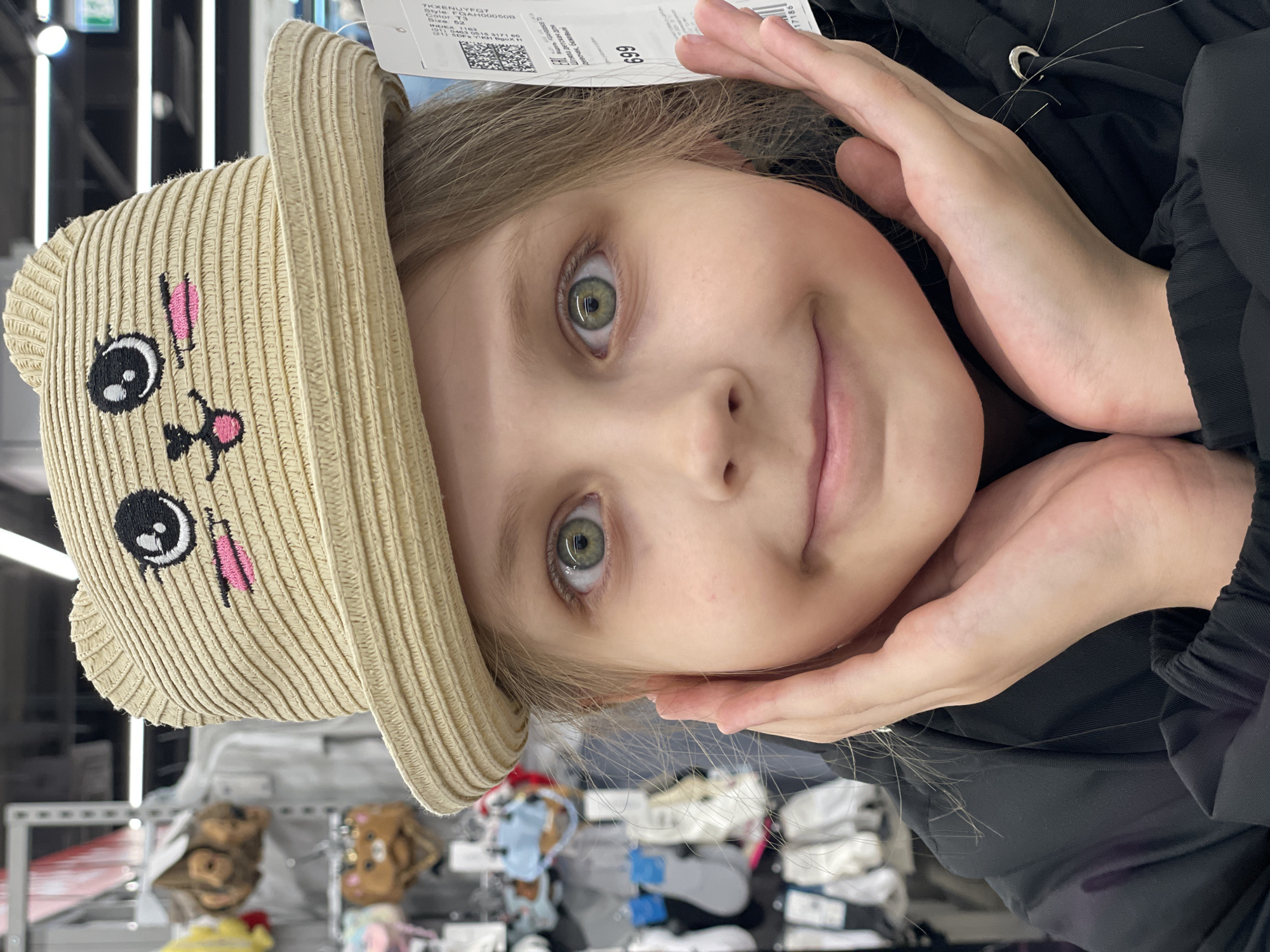 A young girl is seen in a close-up shot, likely in a store. She is wearing a beige sun hat with a cartoon animal face on it, featuring black eyes and a pink nose. Her hands are positioned on either side of her face, framing it. She has fair skin, light blonde hair, and greenish eyes. The background is somewhat blurred and shows shelves with various items, possibly clothing or accessories, indicating the retail setting. The location in Izhevsk, Russia, is not discernible from the image.