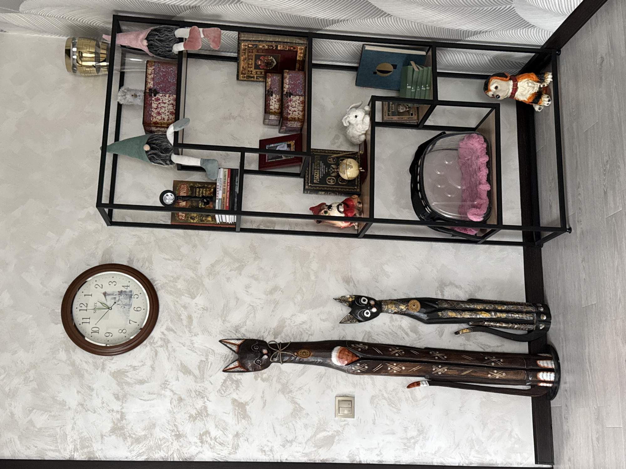 An indoor scene showcases a decorative wall display in Krapivka, Russia. A large black metal open-frame shelving unit is mounted on a light-colored, textured wall. The shelves are adorned with various decorative items, including several gnomes with soft bodies and pointed hats (one in pink, one in green), a small white knitted rabbit, and ceramic figures of a dog and a small cat/fox in a red hat. Numerous books are arranged on the shelves, some with ornate covers in dark red and gold, others in blue and green. Two decorative chest-like boxes with patterned fabric, a small globe, a framed picture, and a clear container holding a pink fluffy item also populate the shelves. A gold-colored lamp fixture is visible near the top left shelf.
Below the shelving unit, a round wall clock with a dark brown frame and a white face showing a landscape picture marks the time at approximately 11:36. The brand "SINIX" is visible on the clock face. To the right of the clock, two long, stylized carved wooden cat figures, dark brown with gold, white, and red embellishments, are mounted on the wall. A light switch is positioned below the wooden cats. No activity or event is taking place in the static display.
Visible text includes: "SINIX" on the clock, and "Гурва" on the spine of one of the books.