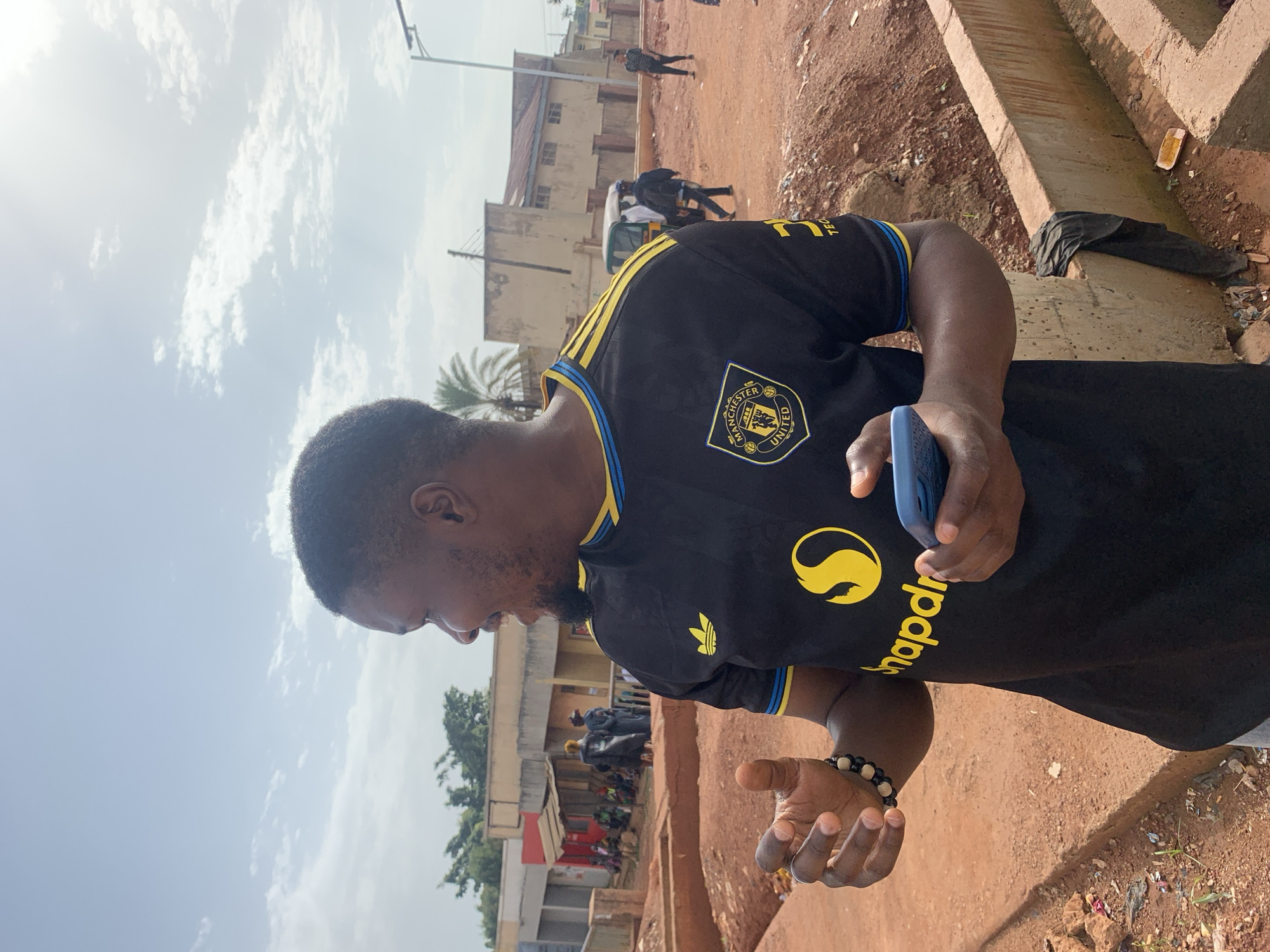 A medium shot captures a Black male in his 20s wearing a black Manchester United jersey with yellow stripes on the sleeves, standing outdoors and looking to his right. He is holding a blue smartphone in his right hand and gesturing with his left hand. A beaded bracelet adorns his left wrist. The background reveals a dusty environment with reddish-brown dirt paths and low concrete structures. In the distance, several buildings with reddish-brown roofs and light-colored walls are visible, along with a few people and what appears to be a three-wheeled vehicle. The sky is partly cloudy. The scene is identified as being in Gura Top, Nigeria.