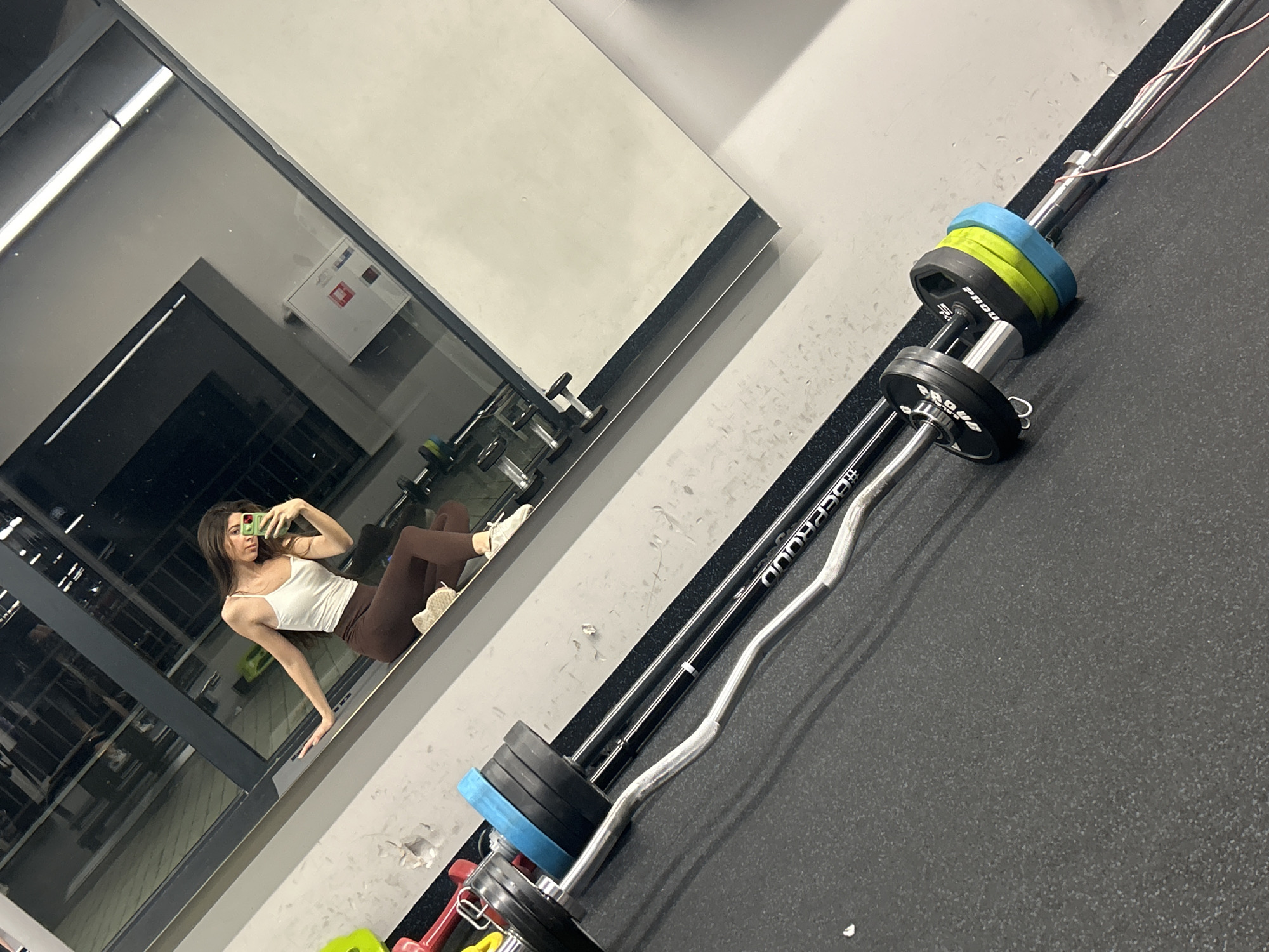 The scene is an indoor fitness gym, likely located in Sofia, Bulgaria. It is captured from a high-angle perspective, showing both gym equipment and a reflection in a large mirror.
In the foreground and right of the frame, a barbell with several weight plates is visible. The barbell is metallic, and the weights are a mix of black, blue, and green. The gym floor is a uniform dark grey.
The left side of the frame showcases a large mirror reflecting a woman taking a selfie. She is wearing a white tank top and brown leggings, and she is holding a smartphone. The mirror also reflects other gym equipment, including dumbbells on racks.
The lighting appears to be artificial, with bright overhead lights. The gym seems clean and well-maintained.