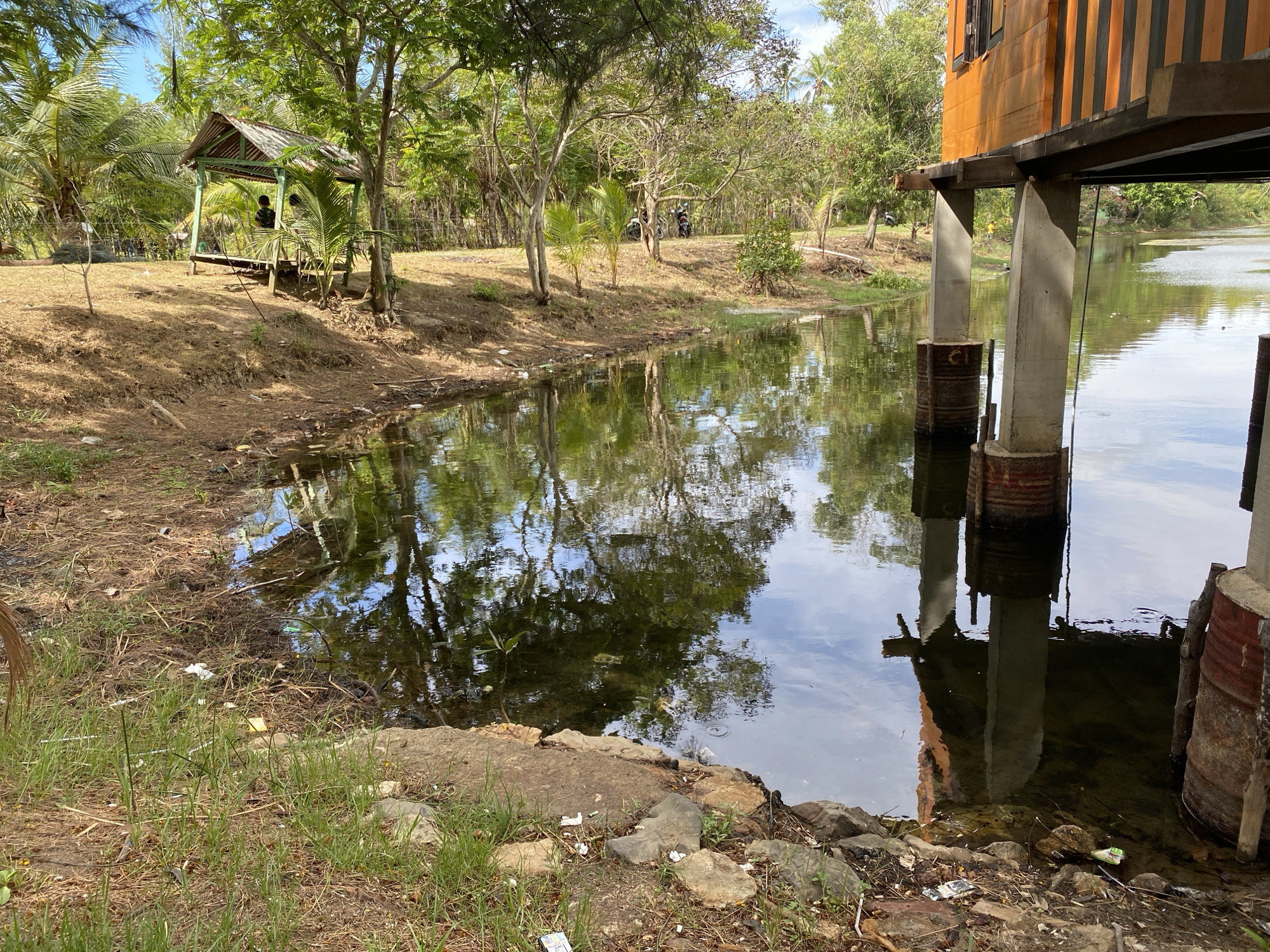 The image depicts a body of water adjacent to a grassy and dry, littered embankment under a bright sky. In the foreground, the embankment is covered with dry grass, scattered rocks, and debris, including plastic wrappers and other refuse. The water's surface reflects the surrounding trees and the sky, creating a mirror-like effect. On the left side of the image, a small, open-sided wooden structure with a thatched roof stands on stilts. Two individuals are visible within or near this structure. Further back, several trees and some palm trees line the embankment. A fence is visible behind the trees. On the right side of the image, a structure built on stilts extends over the water. This structure has orange and brown vertical siding and is supported by concrete pillars, some of which are reinforced with rusted metal barrels. The overall impression is a somewhat neglected waterside environment, possibly in a rural or semi-rural setting.
