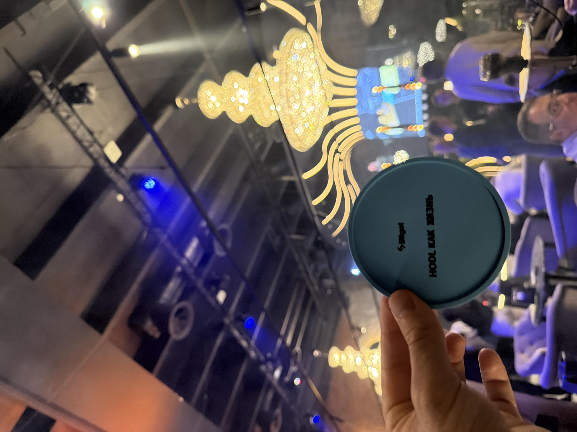 A hand holds a blue coaster with "Bitget" and "HODL KAK ZHIZN" printed on it. The location appears to be a venue with a large, ornate chandelier. In the background, there are blurred figures of people and lighting rigs. The venue's architecture, including ceiling structures and lighting design, suggests it is in Moscow, Russia. The phrase "HODL KAK ZHIZN" is transliterated Russian, which is further evidence of the location.