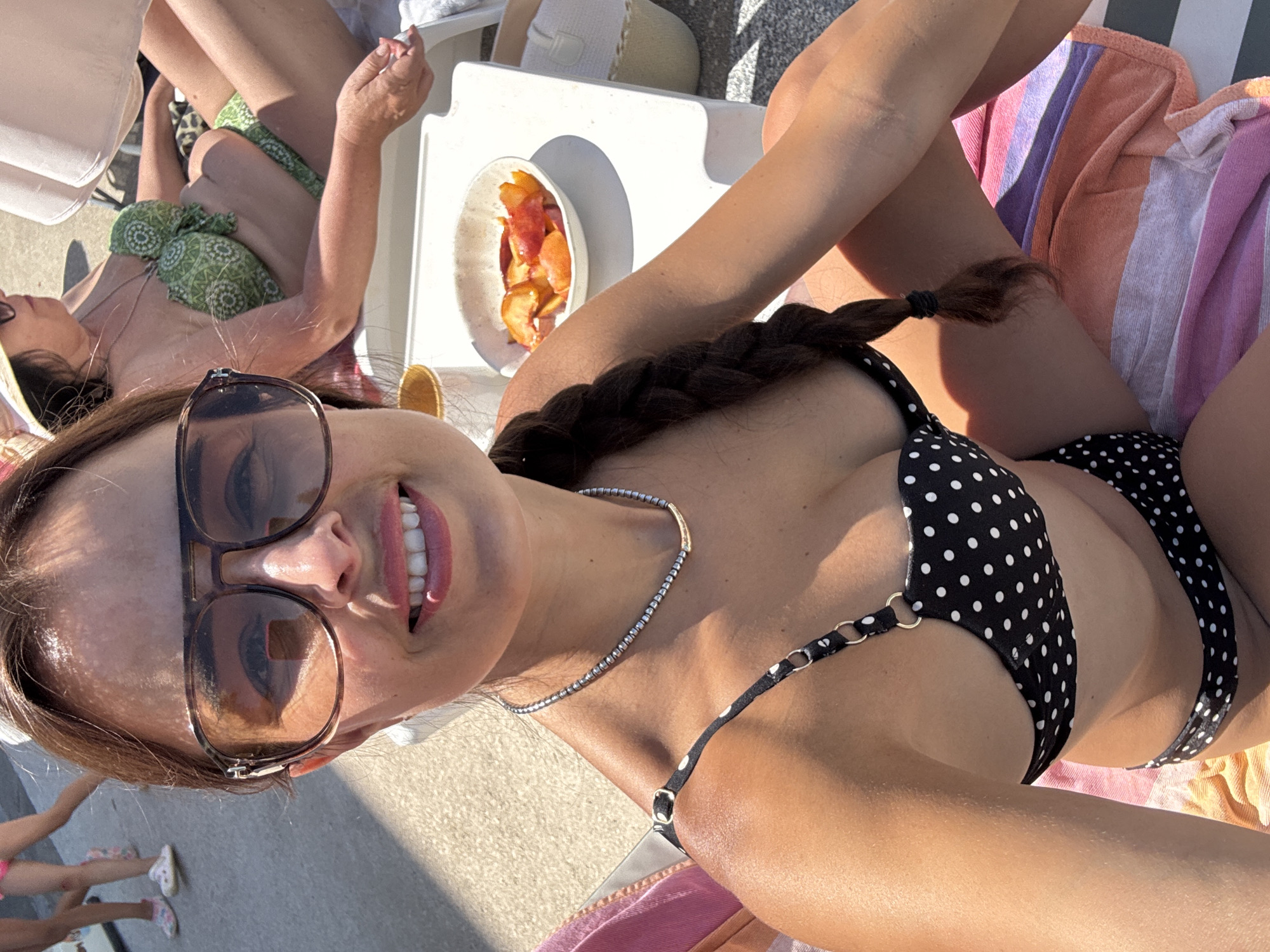 A woman with brown hair, wearing sunglasses and a black polka dot bikini, takes a selfie. She is sitting on a striped towel, with a bowl of sliced fruit on a white table to her right. In the background, two children are walking. Another woman, wearing a green patterned bikini, is reclining on a lounge chair behind the woman in the foreground. The scene appears to be outdoors, possibly at a beach or pool.