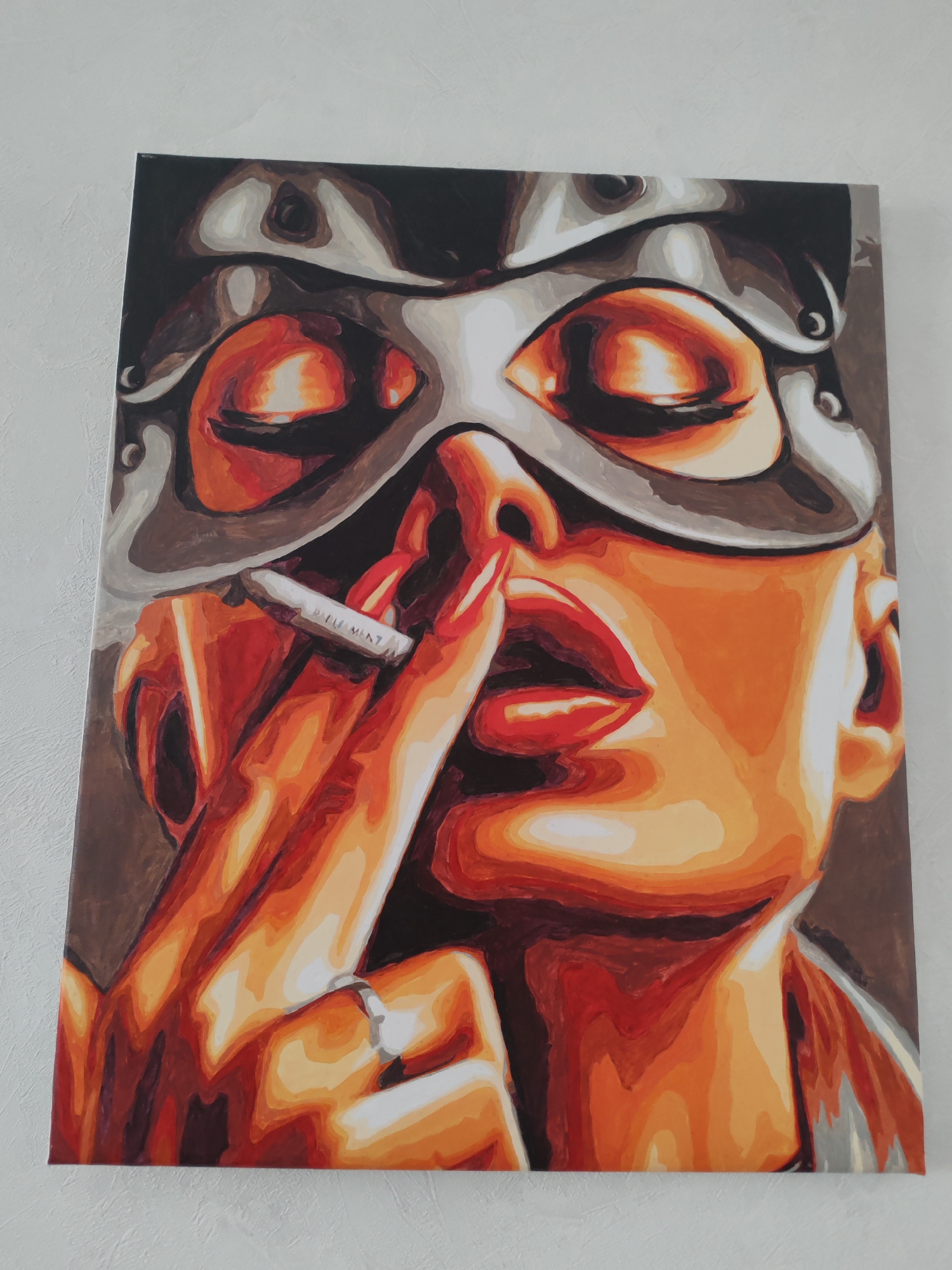 This media file shows a close-up photograph of a painting on a canvas, which is hung on a light-colored, textured wall.
The painting depicts a stylized close-up of a woman's face, shown from the nose down to the upper chest, and her right hand. The woman wears a dark, intricate, metallic-looking mask that covers her forehead and eyes, with her eyes depicted as closed. Her skin tone is rendered in warm shades of orange, red, and brown, with strong highlights and shadows creating depth. Her lips are full and painted bright red, slightly parted to hold a cigarette.
Her right hand, with slender fingers and a ring on the ring finger, is raised to her face. Her index finger is positioned near her lips, touching the cigarette. The cigarette is white with a filter and has the word "ELEMENT" visible on its side. The background of the painting is a dark, indeterminate color, focusing attention on the figure. The art style features bold outlines and distinct blocks of color, giving it a graphic or pop-art aesthetic.