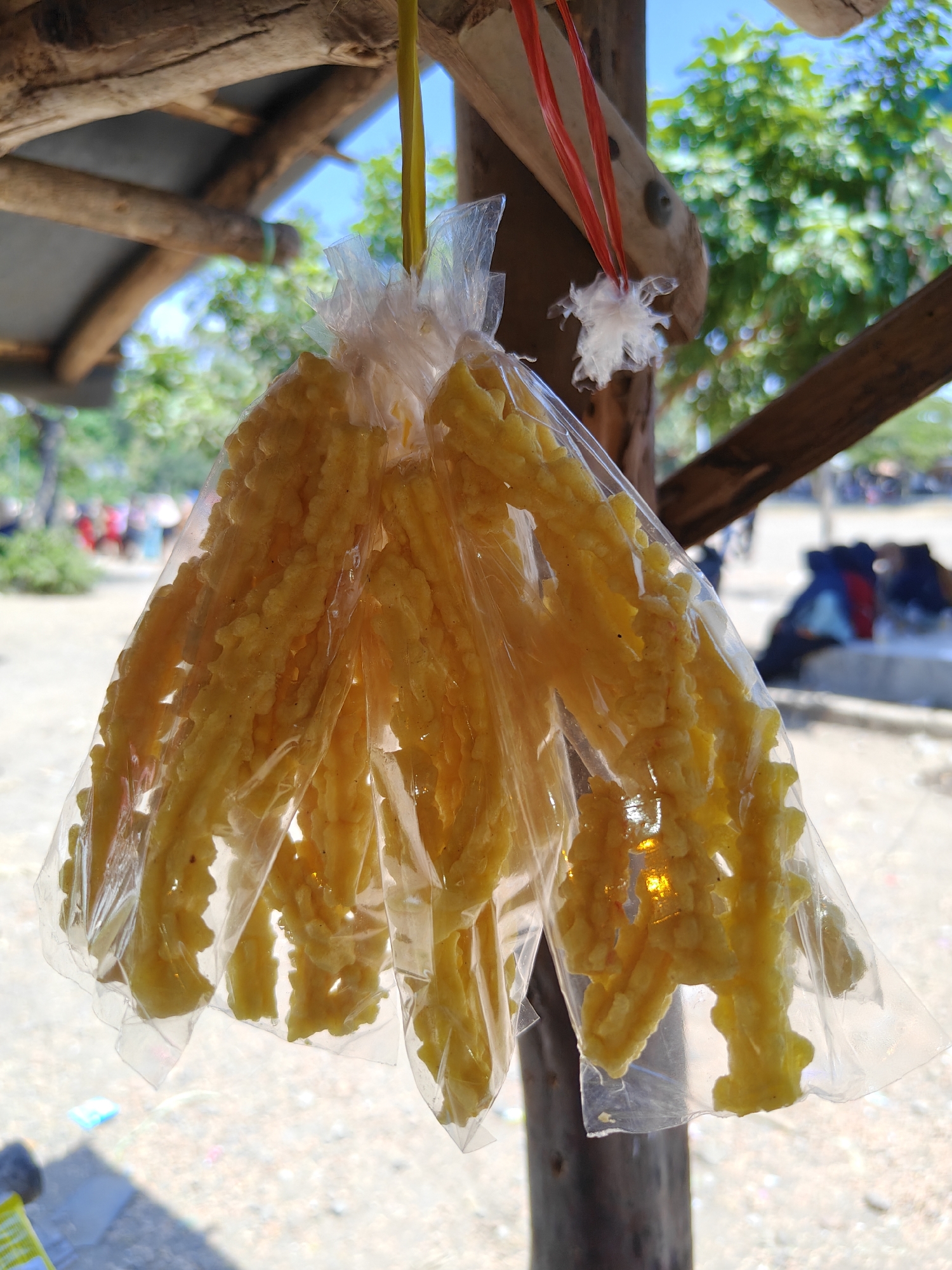 A clear plastic bag filled with bright yellow, intricately textured snack sticks hangs from a yellow string and a red string tied to a wooden beam. The snack sticks have a ridged, almost braided appearance. The background is blurred, showing green foliage, a bright blue sky, and what appears to be an outdoor marketplace. Several people are visible in the distance, some seated and others standing. The ground is covered with light-colored gravel and sand. The overall scene suggests an outdoor vendor stall in Cermee, Indonesia, likely in a tropical or subtropical climate given the lush greenery and bright sunlight.