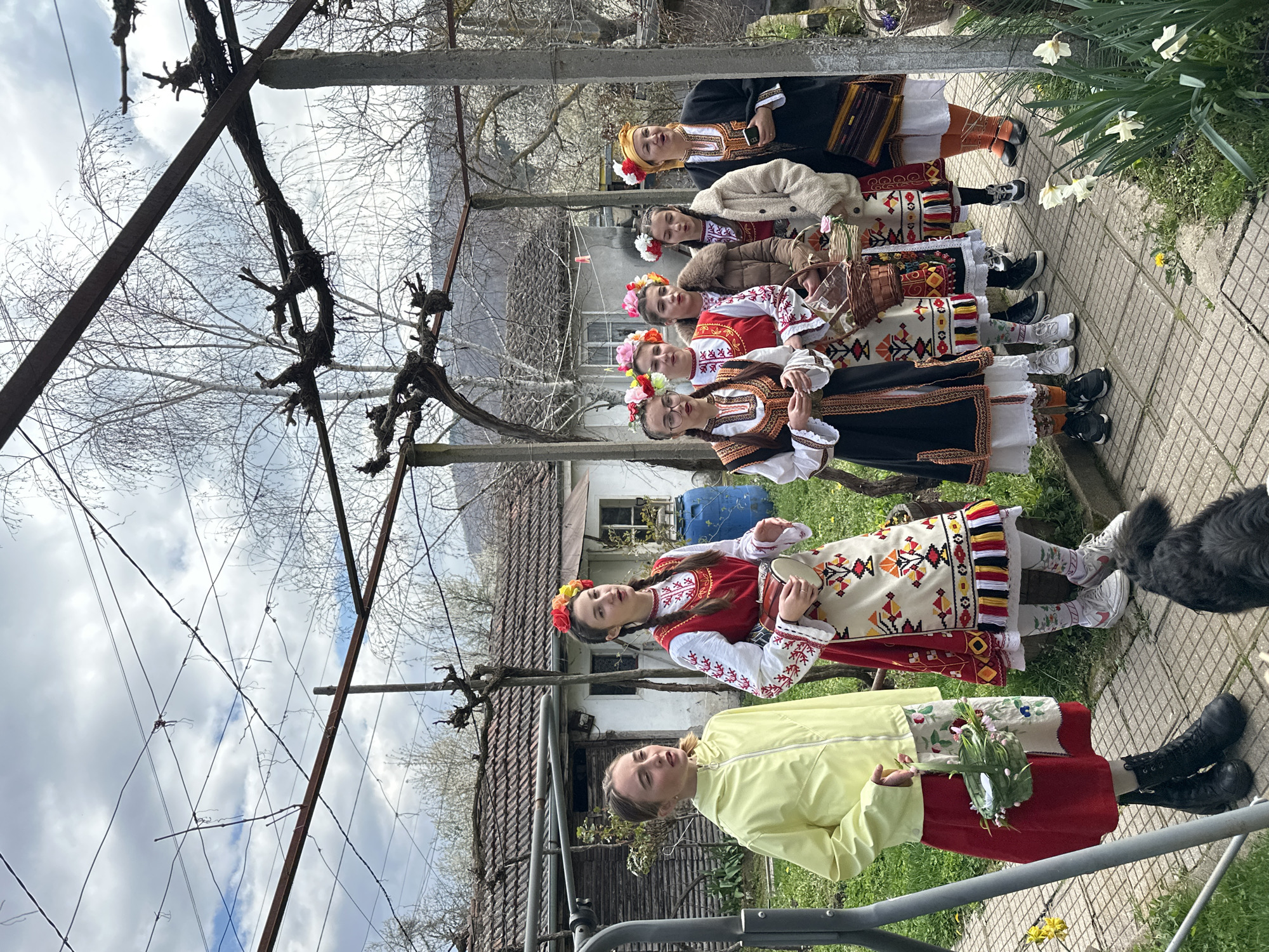 A medium shot, outdoors, shows six women and one girl standing in a line on a paver path. The women are wearing traditional Bulgarian clothing, with white, red, black, and multicolored embroidery. Each has a flower crown on her head. The girl stands at the front of the line, dressed in a yellow jacket and red skirt, holding a bouquet of flowers.
Behind them is a rural scene: a white house with a brown roof, trees, and mountains in the distance. An arbor with bare vines is visible on the left. Daffodils grow on the right side of the path.
The location is likely Mrachenik, Bulgaria, as requested.