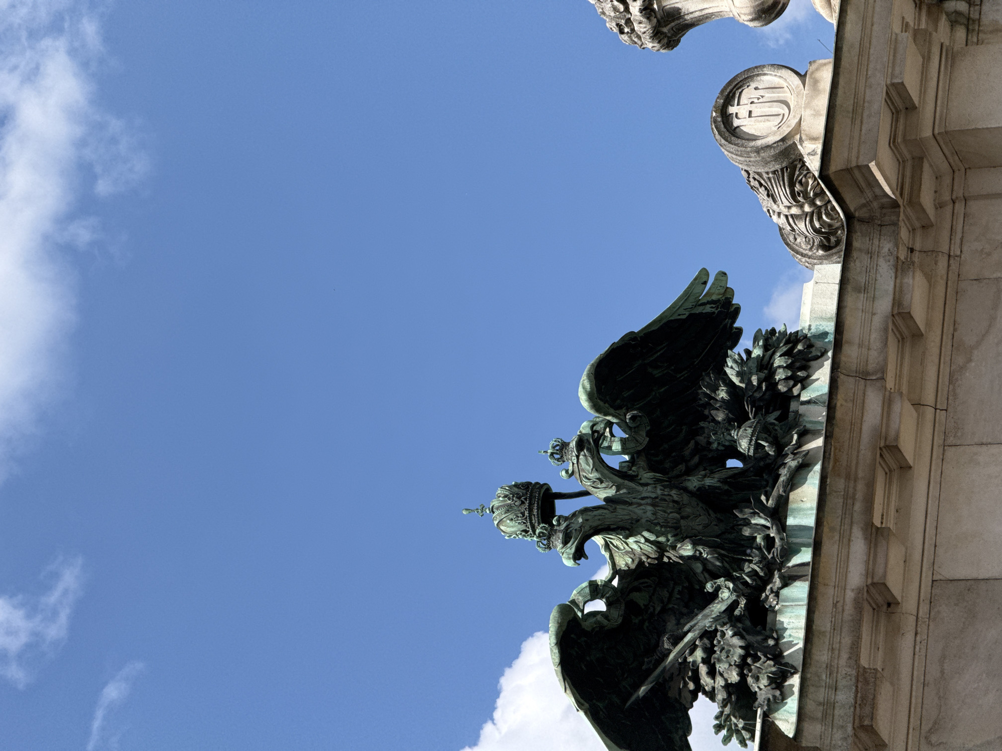 This upward-angled photograph captures a majestic and weathered bronze sculpture of a double-headed eagle, adorned with crowns, set against a bright blue sky with scattered white clouds. The eagle, a historical symbol often associated with the former Austro-Hungarian Empire, is intricately detailed with feathers and appears to be part of a grand architectural embellishment, consistent with a significant historical building in Vienna, Austria. The eagle is surrounded by a bronze laurel wreath or similar foliage.
To the right of the eagle, part of the stone building's facade is visible, showcasing ornate architectural details. These include a carved stone corbel or bracket with intricate scrollwork and foliage. On a circular emblem within this carving, a distinct stylized letter or symbol, possibly an "S" or a monogram, is discernible. Further architectural elements and stone blockwork are visible, suggesting a classical or imperial architectural style. The bright, clear daylight indicates it is daytime with fair weather. No specific activity or human presence is depicted, as the image focuses on the static sculpture and architecture, conveying a sense of historical grandeur and imperial legacy.
