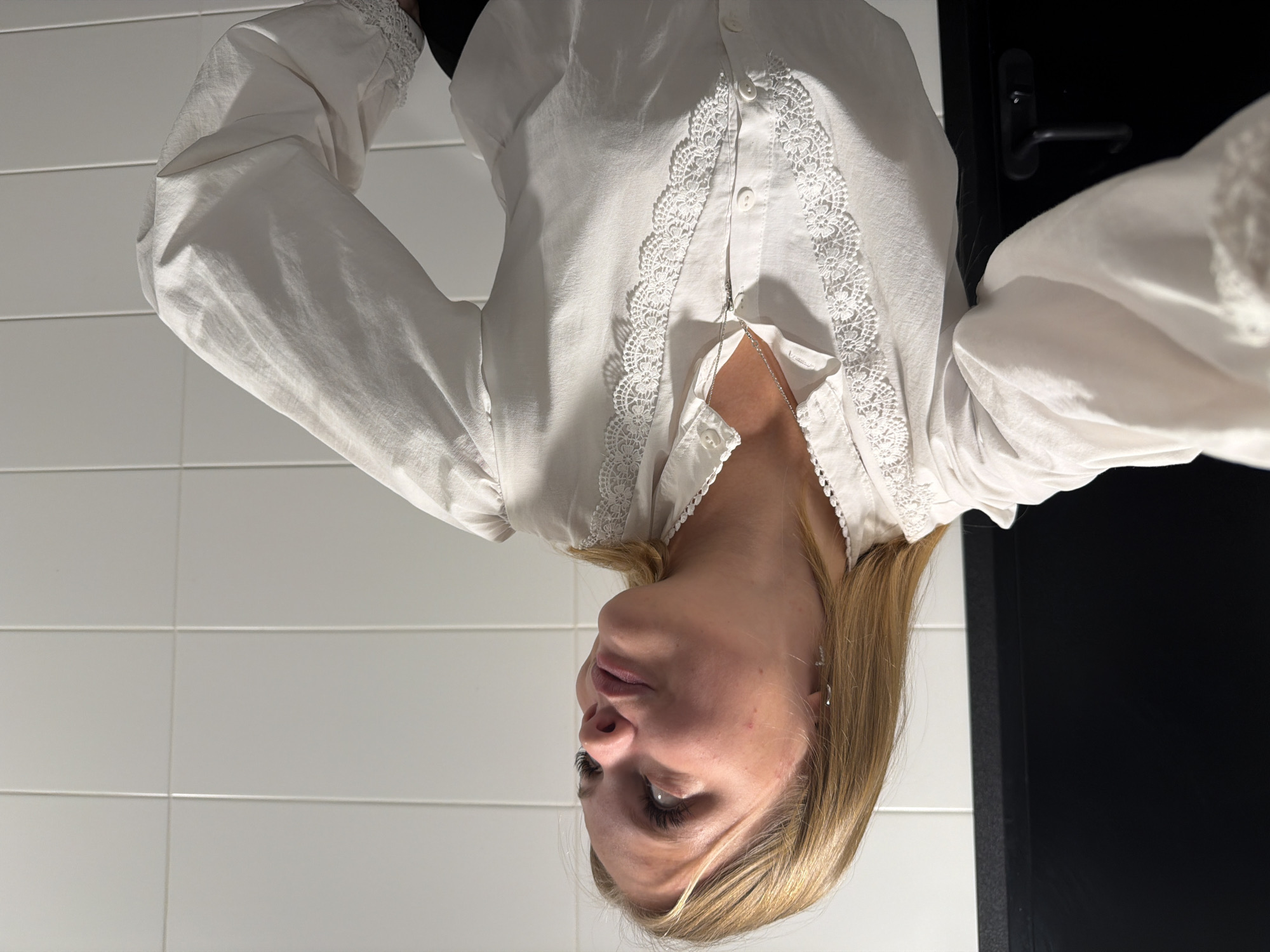 A young Caucasian woman with blonde hair is captured in a medium close-up, taken indoors. She is wearing a white blouse adorned with intricate lace detailing around the collar and down the front placket. A delicate silver necklace with a small pendant rests against her chest. Her gaze is directed upwards and to her right, with a slight pout to her lips. She has false eyelashes. The background features a black door on the left and white tiled walls on the right, suggesting a bathroom or changing room environment. The lighting is bright, casting subtle shadows. This scene is not specifically identifiable as being in Izhevsk, Russia, as the image lacks any distinguishing geographical markers.