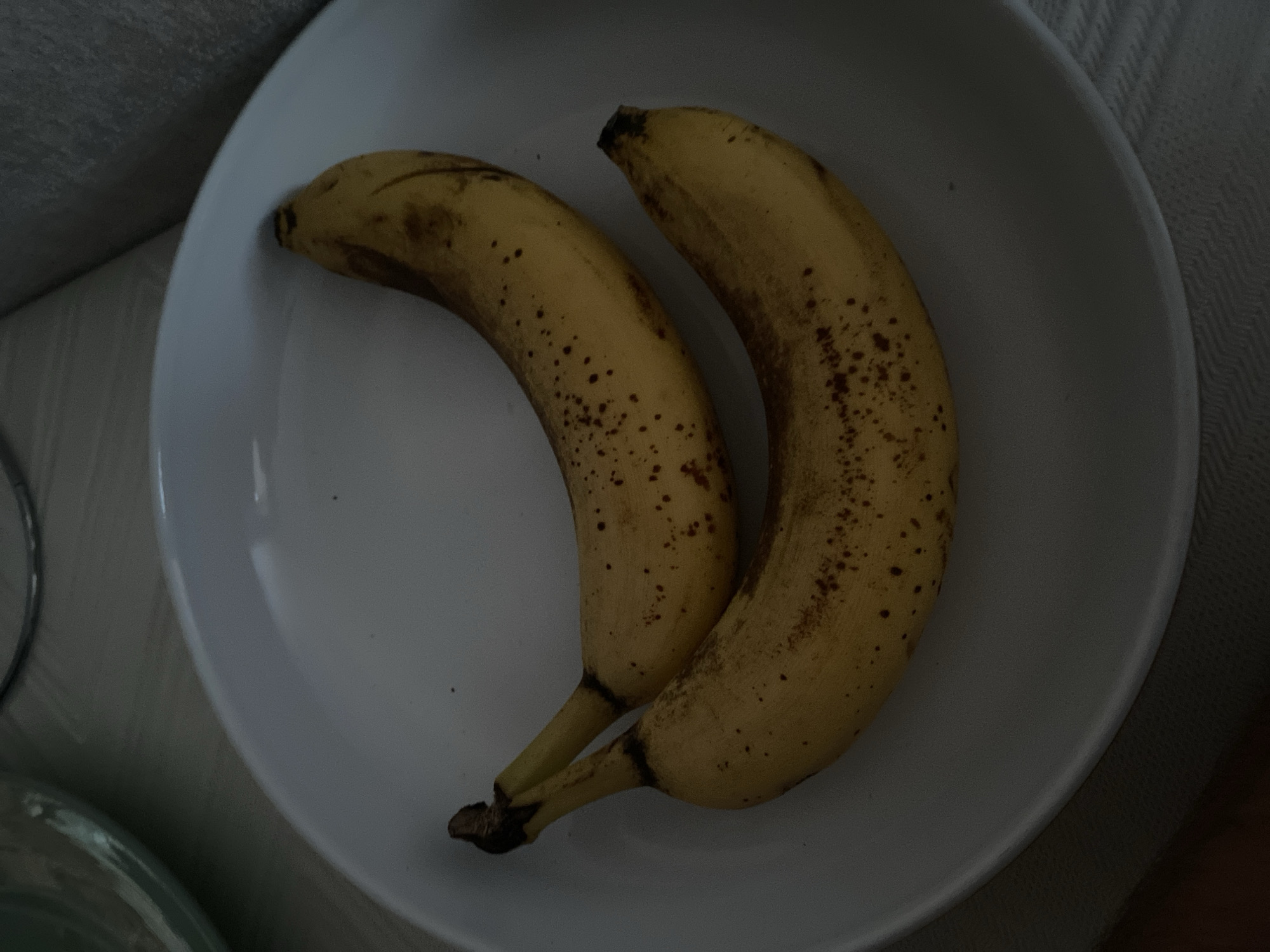 Two ripe bananas with brown spots are arranged diagonally on a white plate. The plate is centered on a light-colored, textured surface, possibly a tablecloth or placemat. A portion of a light green bowl or dish is visible in the upper left corner of the image. The background consists of a blurred, gray surface, likely furniture. The scene appears to be indoors, possibly in a kitchen or dining area. There is no indication of the location being in Chișinău, Moldova.
