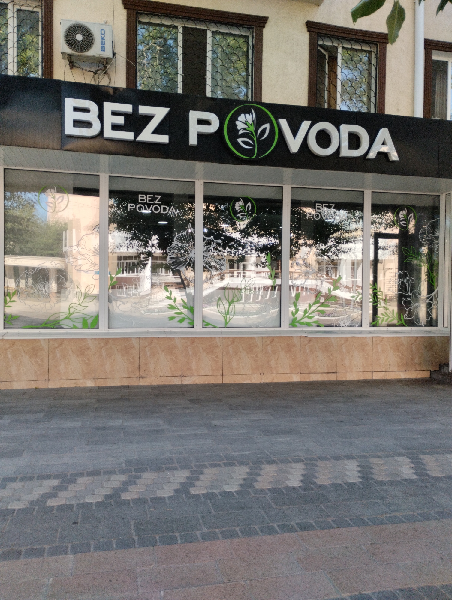 The image shows a store exterior in Kostanay, Kazakhstan. The store's name, "BEZ POVODA", is displayed in large white letters on a black background above the storefront windows. The windows are decorated with white and green floral and leaf patterns. The building has a light-colored facade with brown trim around the windows. An air conditioning unit is visible mounted on the wall above the store. The pavement in front of the store is made of gray and tan interlocking bricks, with a decorative pattern in the center. The overall scene depicts a commercial storefront on a sunny day.