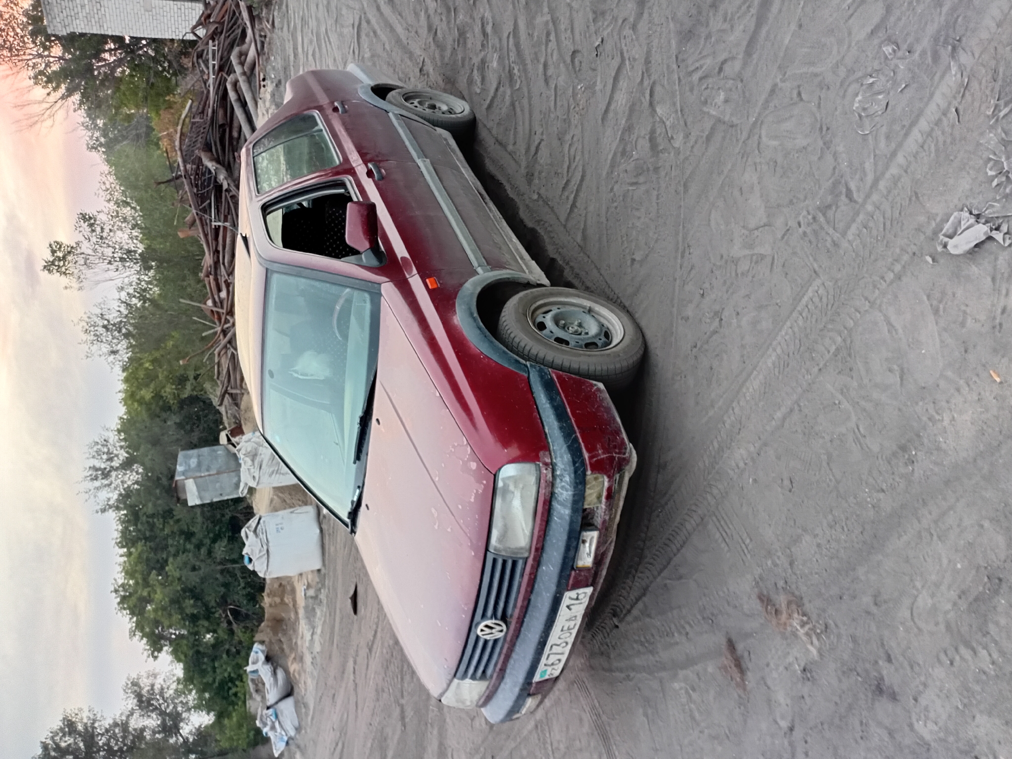 An aged, dark red Volkswagen Jetta sedan, with visible dirt and wear, is parked on a dusty, unpaved lot. The car has its driver's side window open. In the background, a pile of cut lumber and debris is visible, along with several large white sacks, possibly containing building materials, and a small, metallic shed. The environment appears to be an outdoor storage or construction area, likely on the outskirts of Semey, Kazakhstan, given the license plate on the car. The sky above is overcast with hints of sunset colors. The ground is mostly dirt with tire tracks from vehicles.